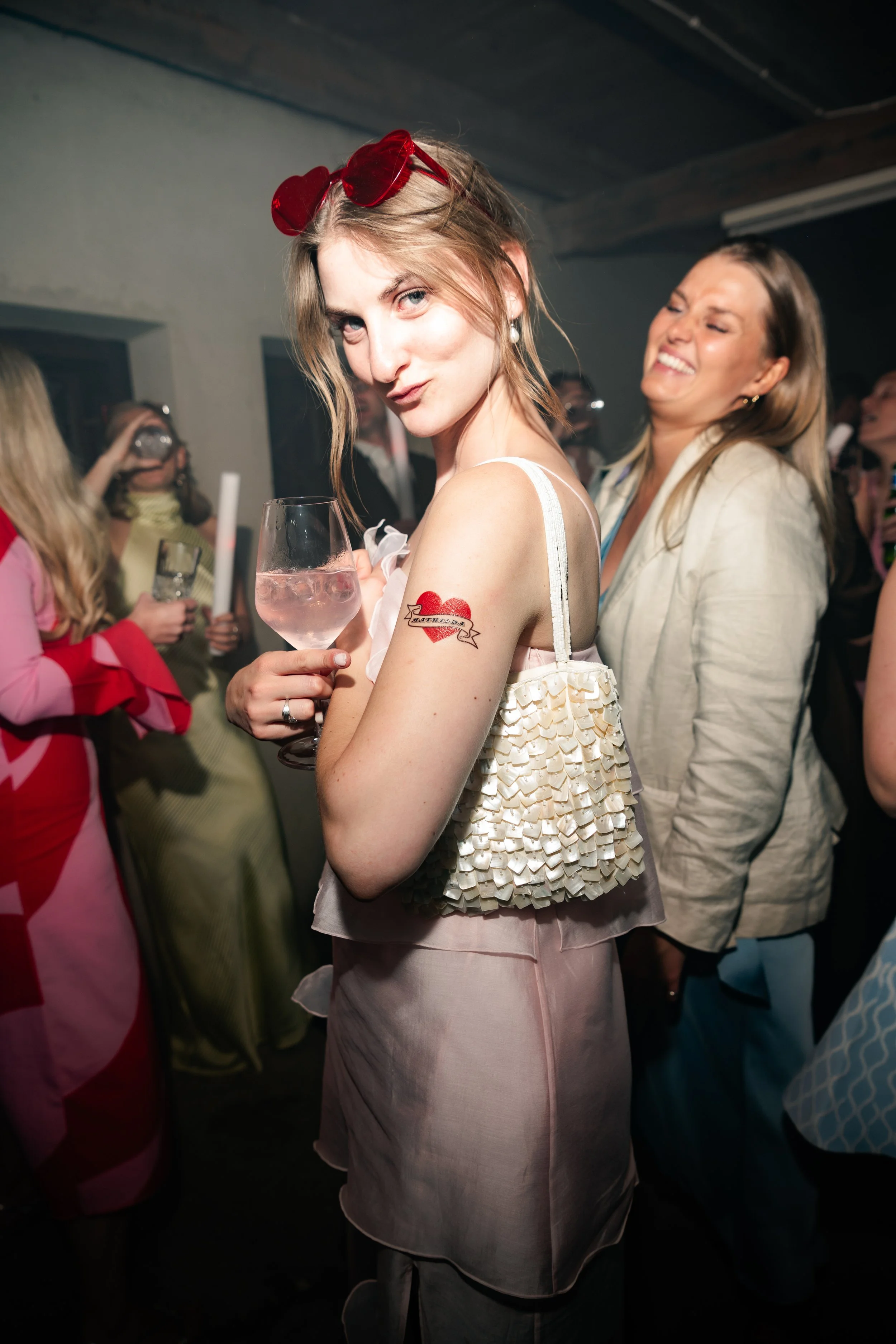 A young woman with red sunglasses on her head, holding a glass of pink drink, poses at a party with a tattoo of a red heart on her arm. She is wearing a sleeveless top with decorative fabric and has a playful expression.