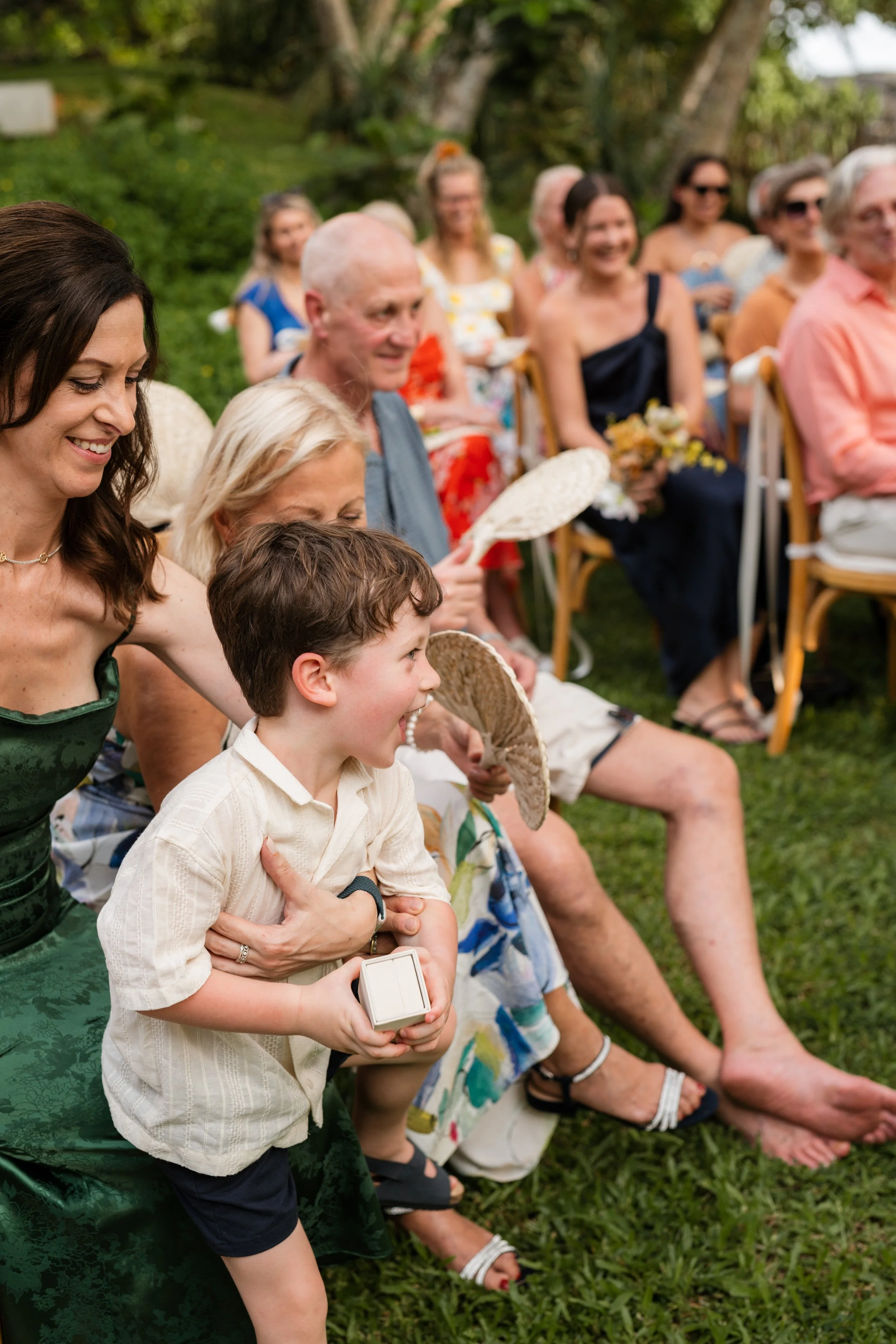 A group of people attending an outdoor event, with some women and children seated on chairs outdoors surrounded by greenery, smiling and engaging in the event.