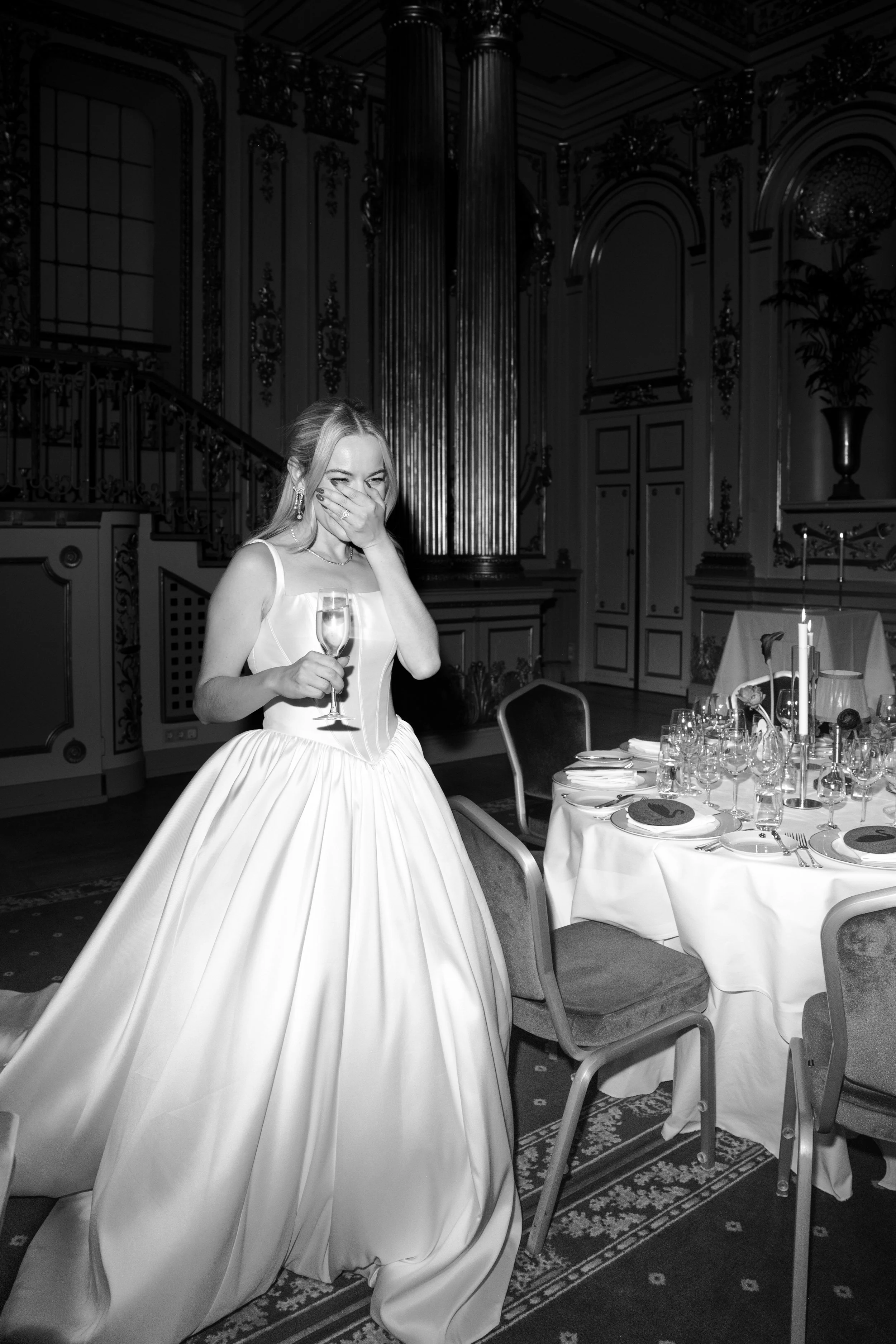 A woman in a wedding dress holding a glass of champagne, laughing and covering her mouth, at a wedding reception in an elegant, ornately decorated room with a grand pipe organ in the background and a table set for dinner.