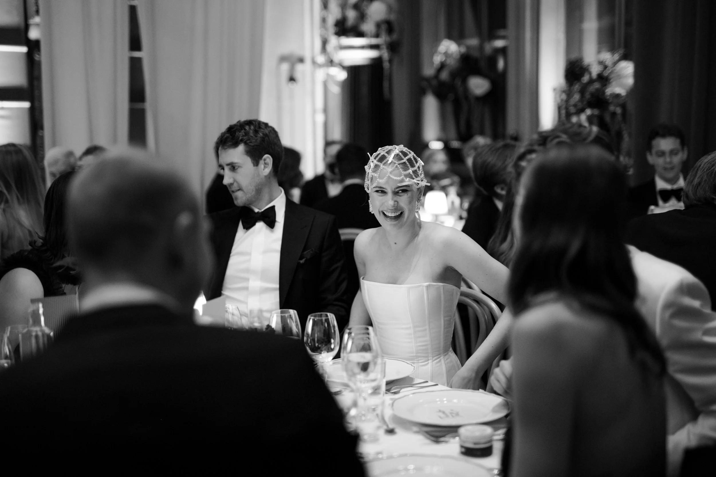 A woman in a strapless dress and headpiece laughing at a formal dinner event, surrounded by guests in tuxedos.