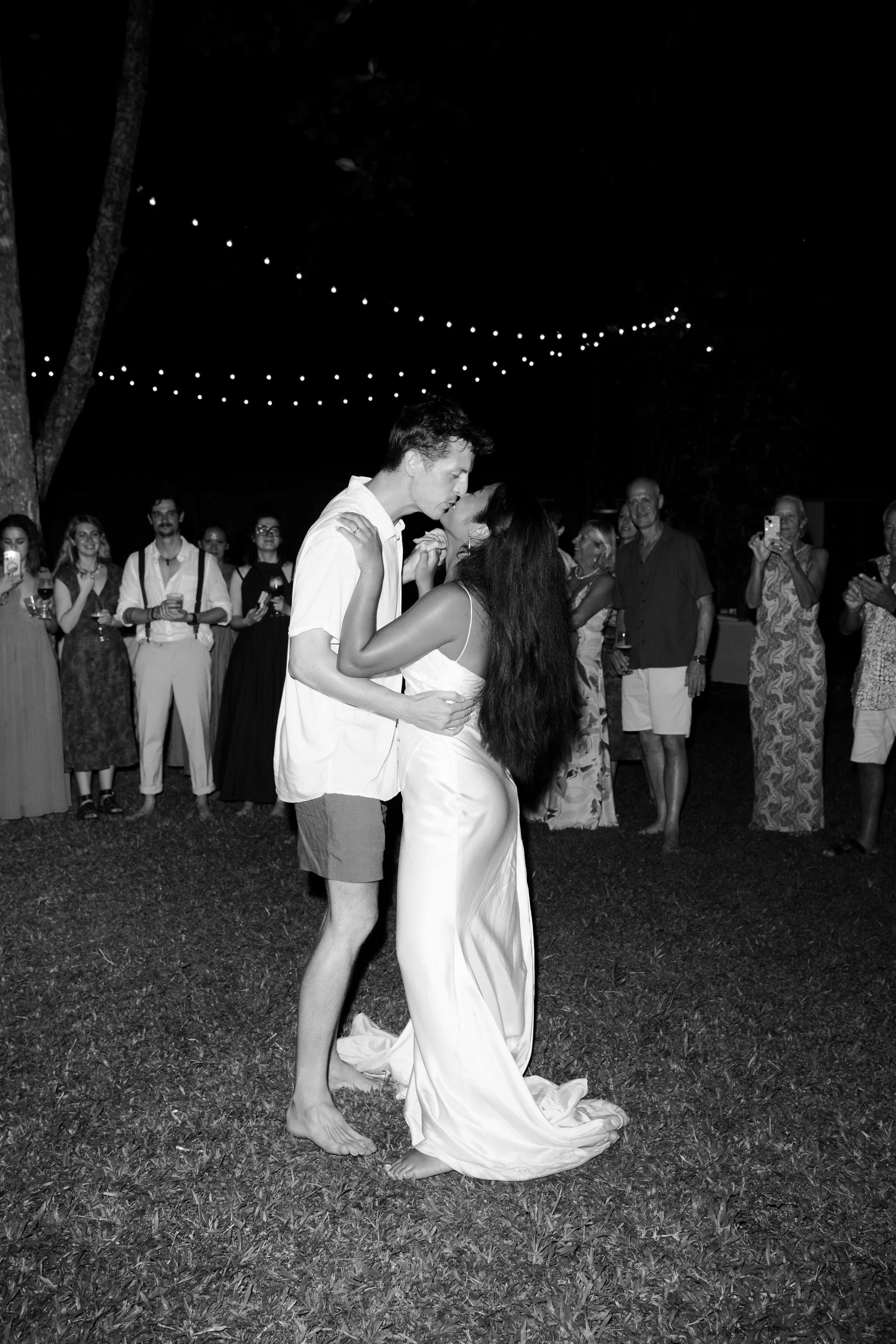 A couple sharing a kiss during their wedding dance at night, surrounded by guests under string lights.