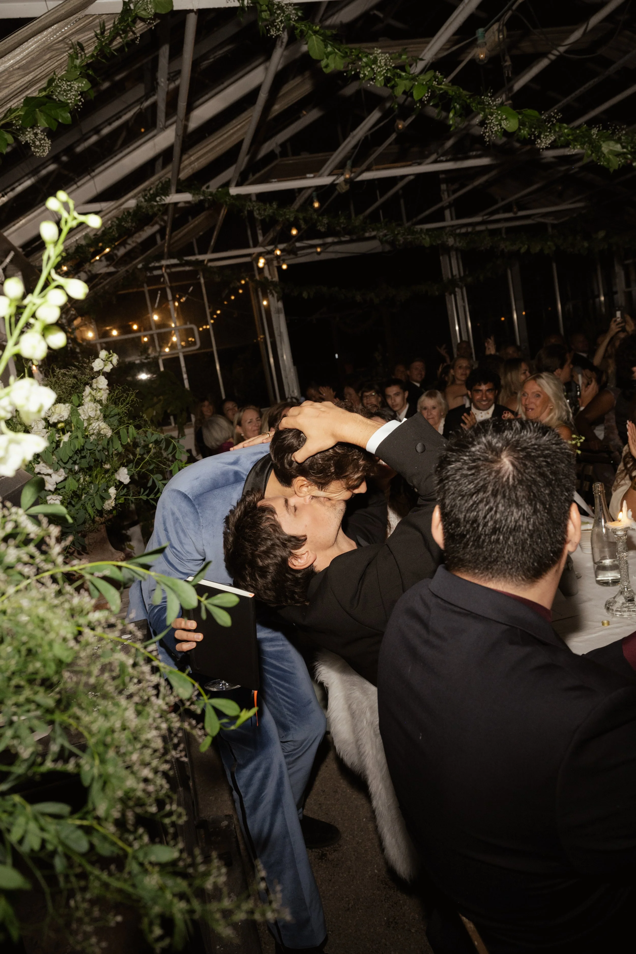 Two men are kissing and embracing at a wedding reception with guests seated at long tables, decorated with flowers and greenery, inside a glass-walled venue at night.