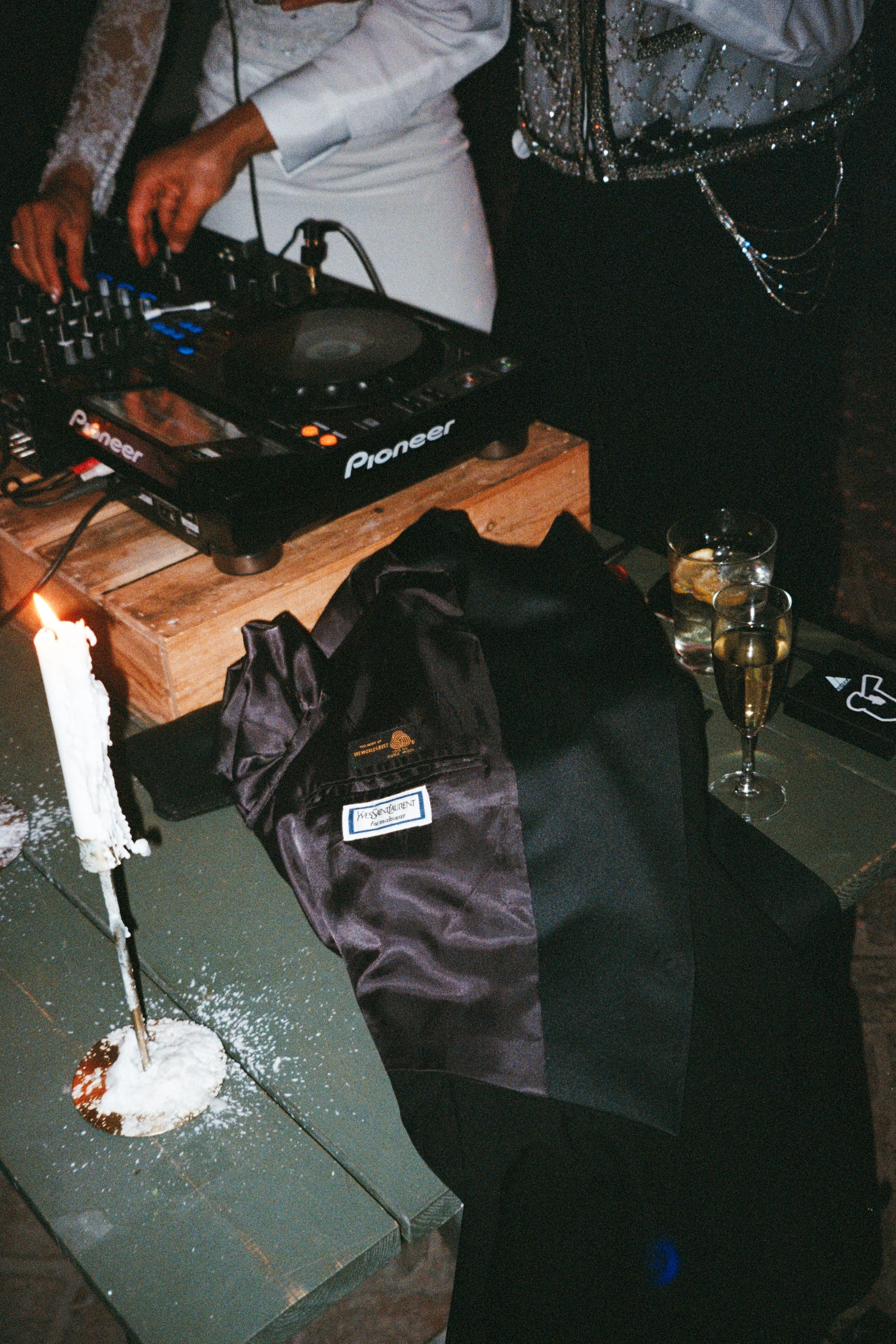 Close-up of a DJ's turntable setup on a wooden table, with a person adjusting controls, and two glasses of champagne nearby, in a dimly-lit environment.