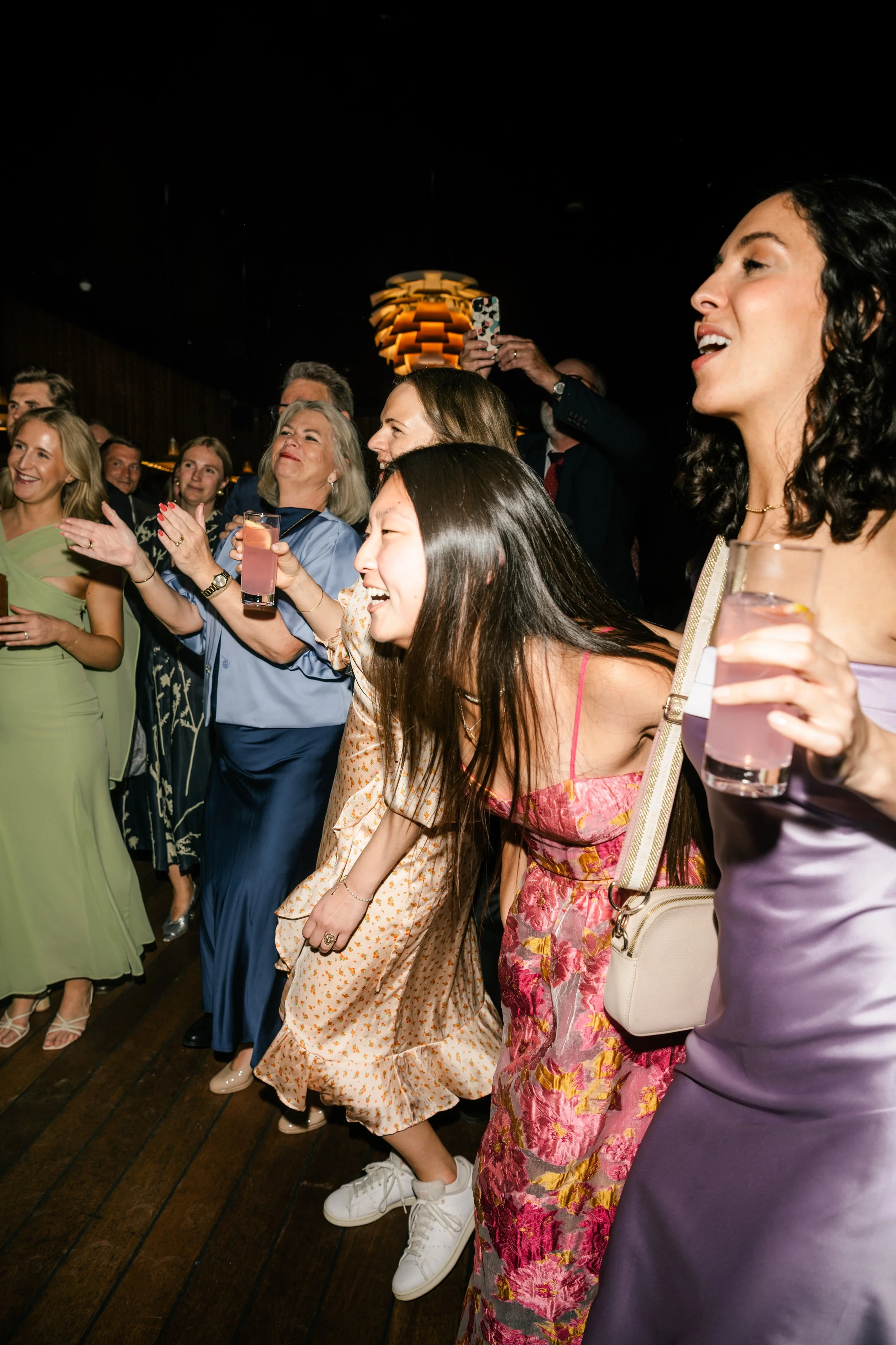 Group of women dancing and having fun at a party, some holding drinks, in an indoor venue with a dark background and modern lighting.