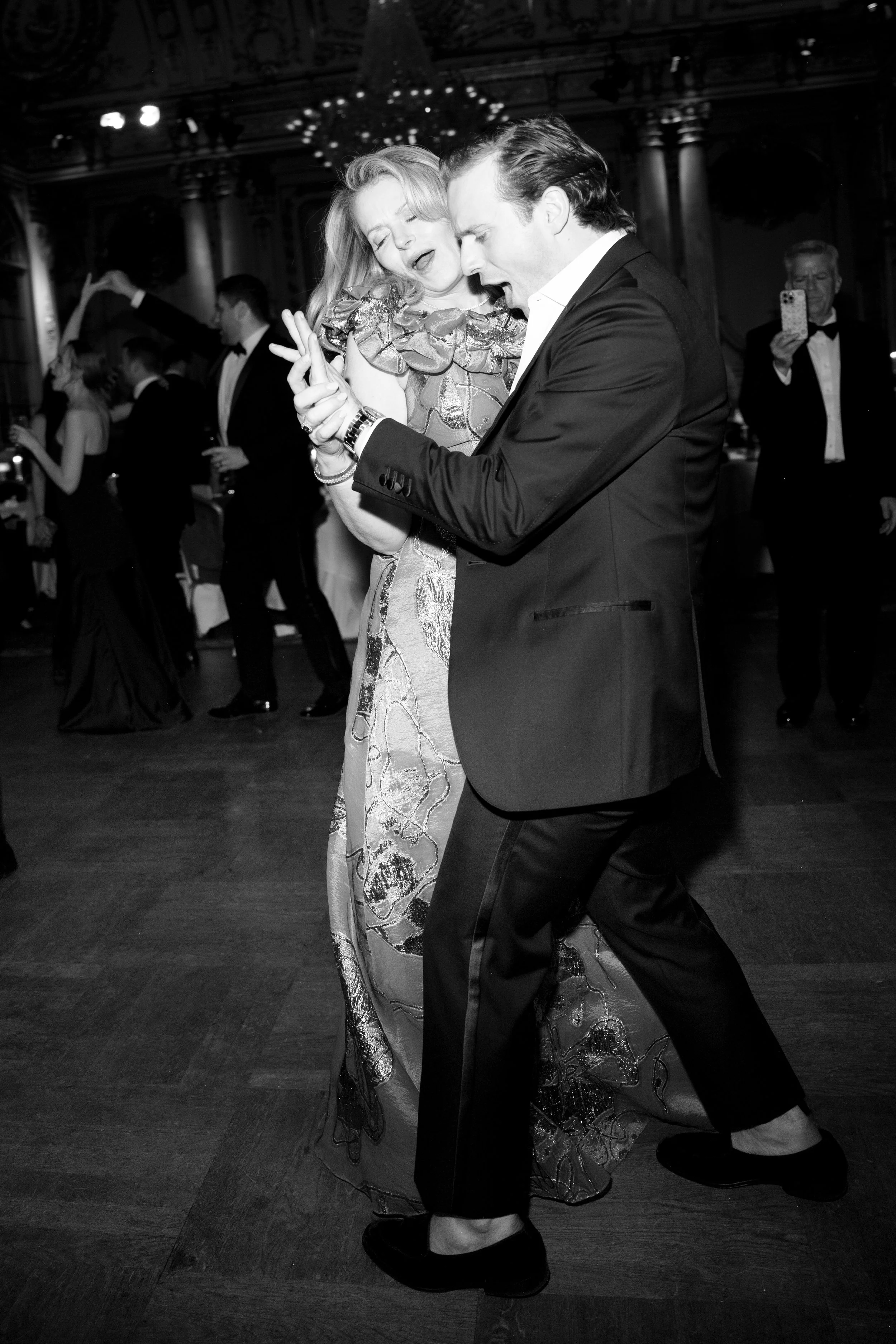 A couple dancing closely at a formal event, with other guests dancing and taking photos in the background.