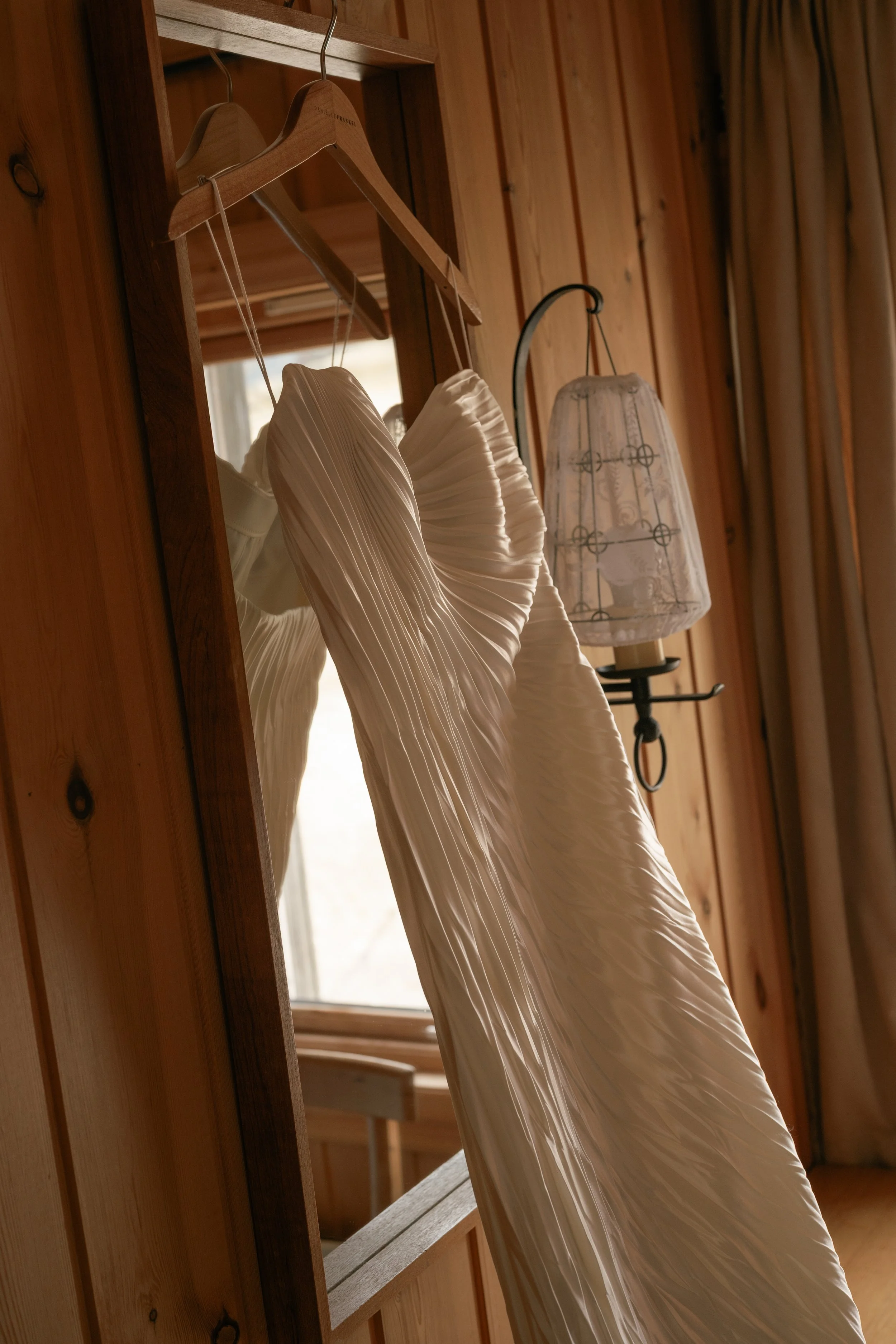 A white wedding dress hanging on a wooden hanger in front of a mirror, with a window and wooden paneling in the background.