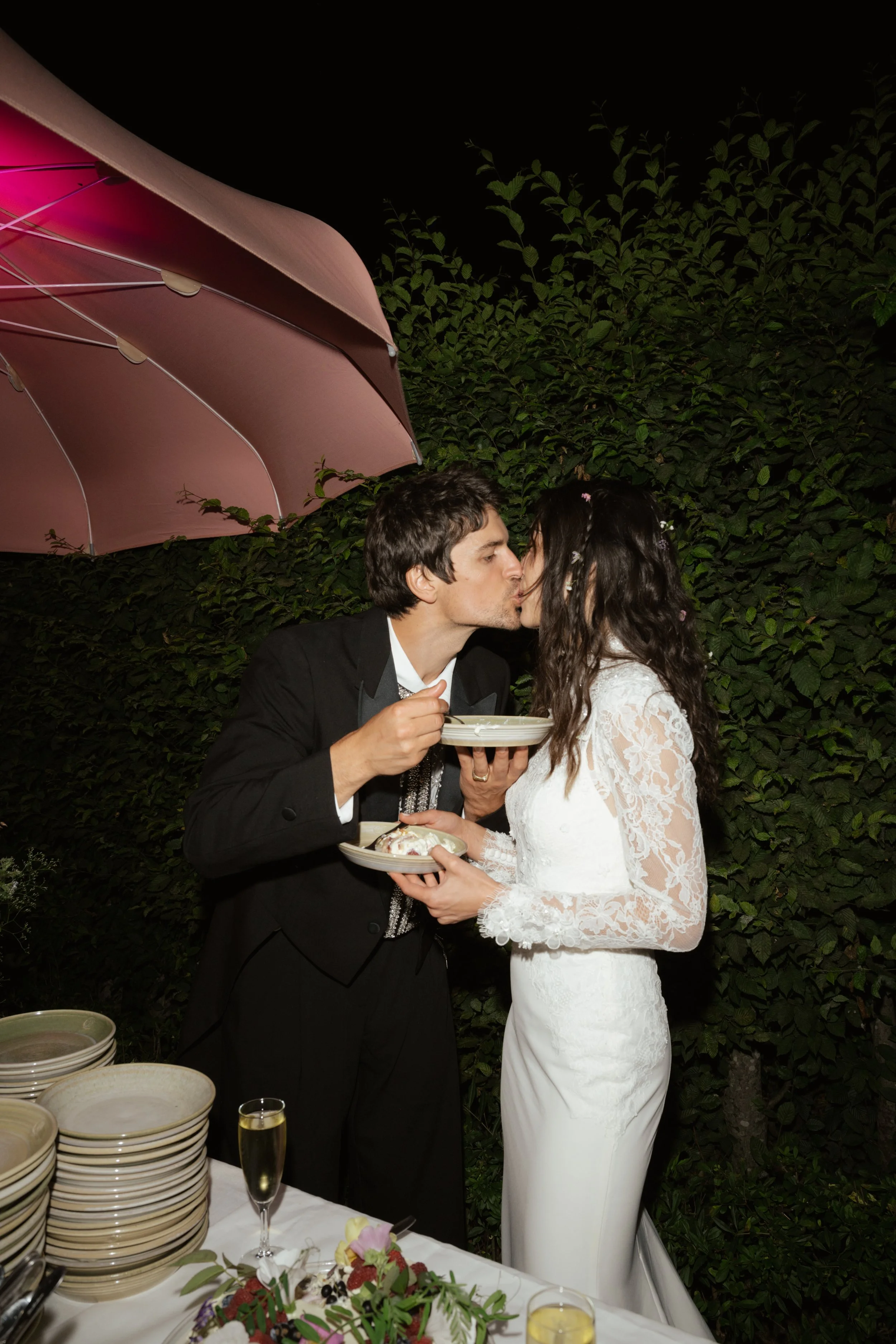 A newlywed couple sharing a kiss under a pink umbrella at night, with a table of food and drinks in front of them.