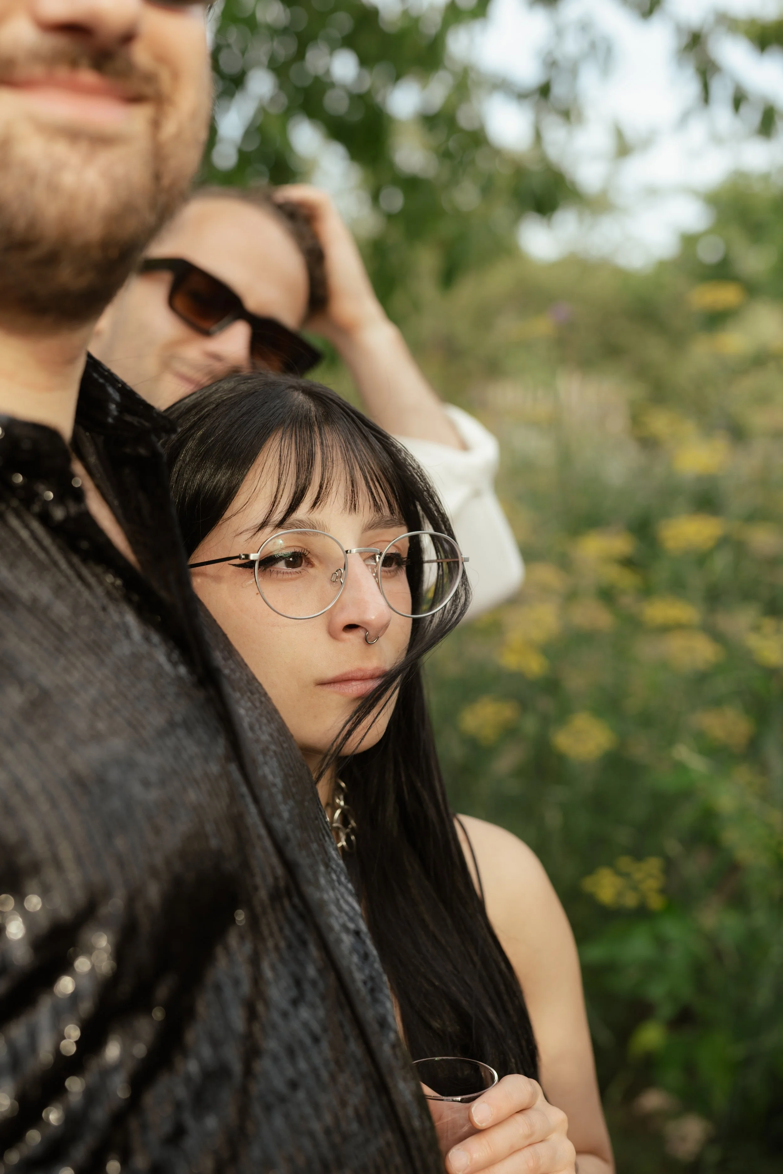 A group of friends standing outdoors with blurred trees in the background, with one woman in focus wearing glasses and a septum piercing.