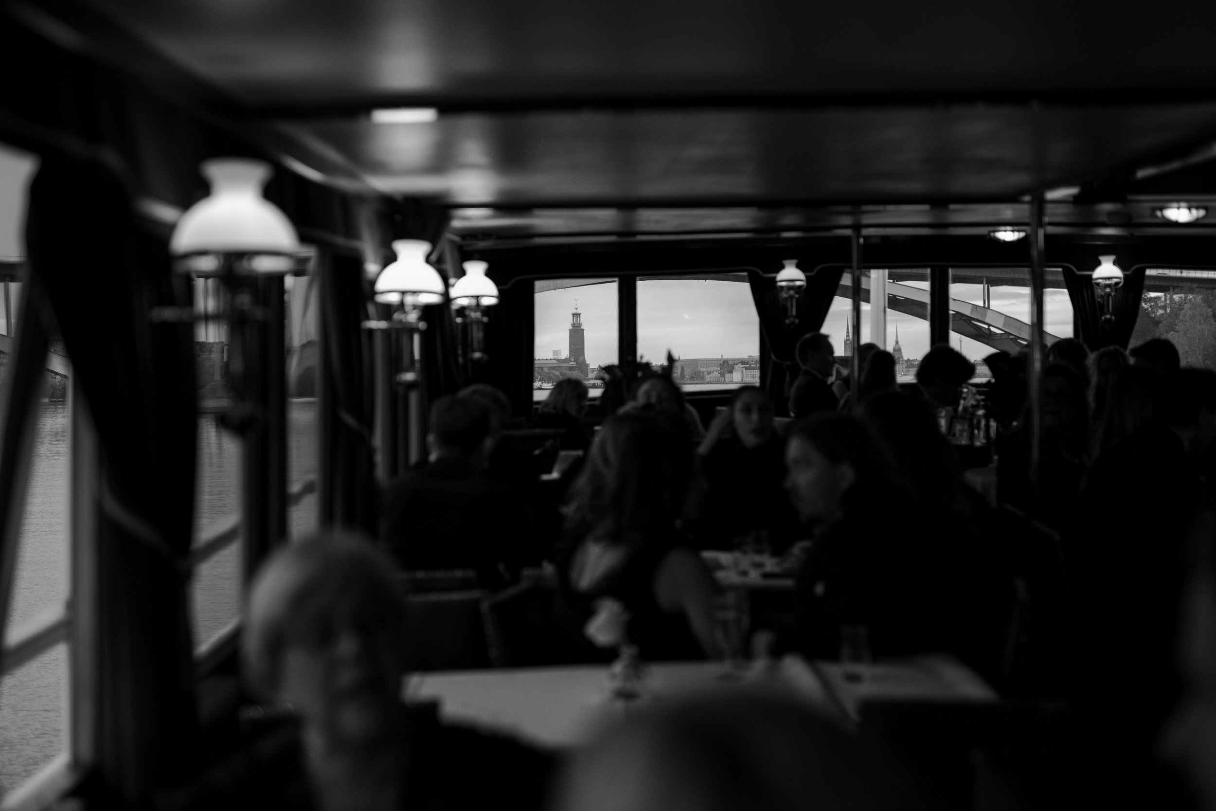 The interior of a boat with passengers dining, with a view of buildings and a tower through the window.