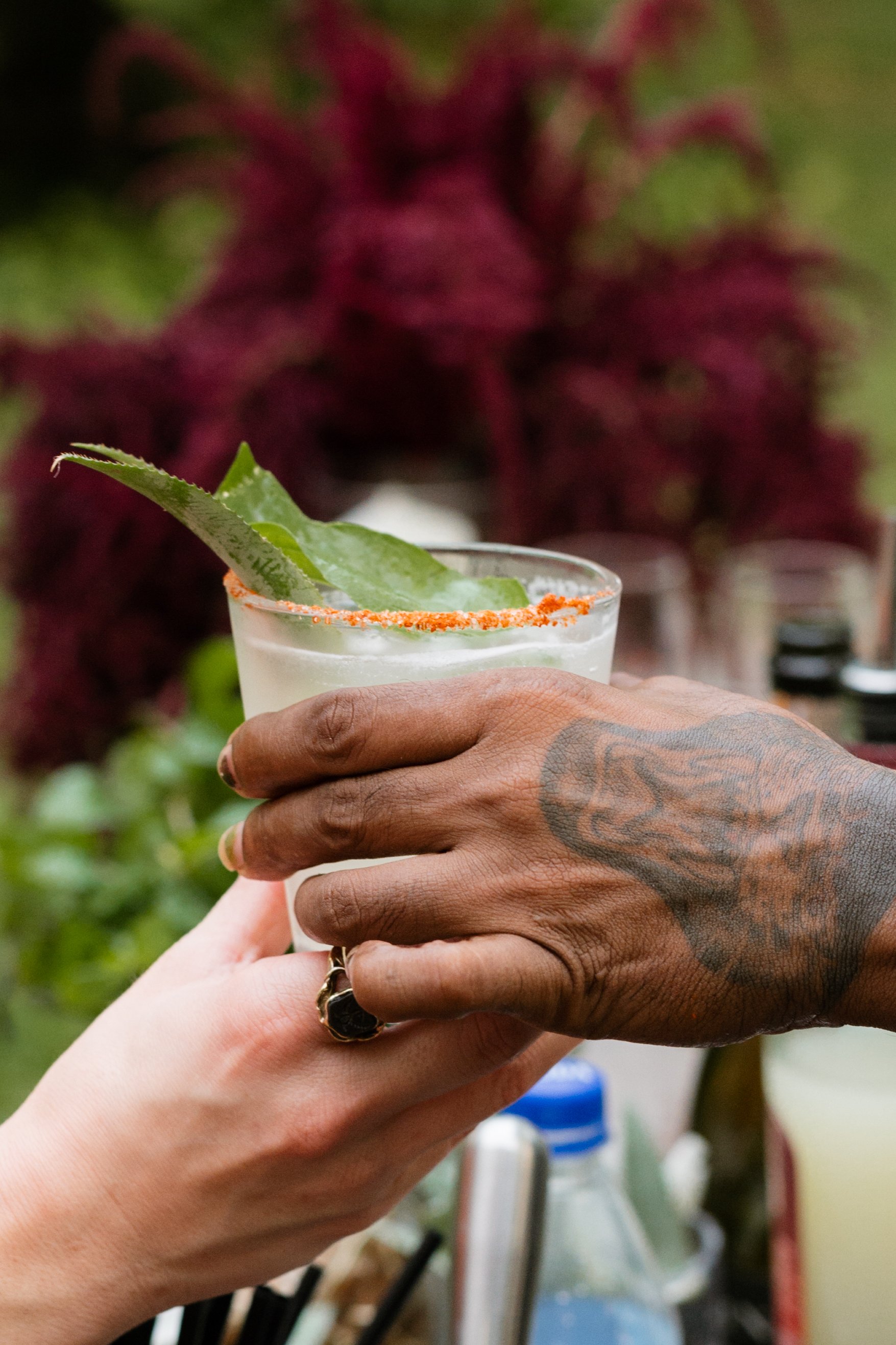 Two hands, one with tattoos, holding a drink with a salted rim and a lime slice, with a blurred greenery and purple plants in the background.