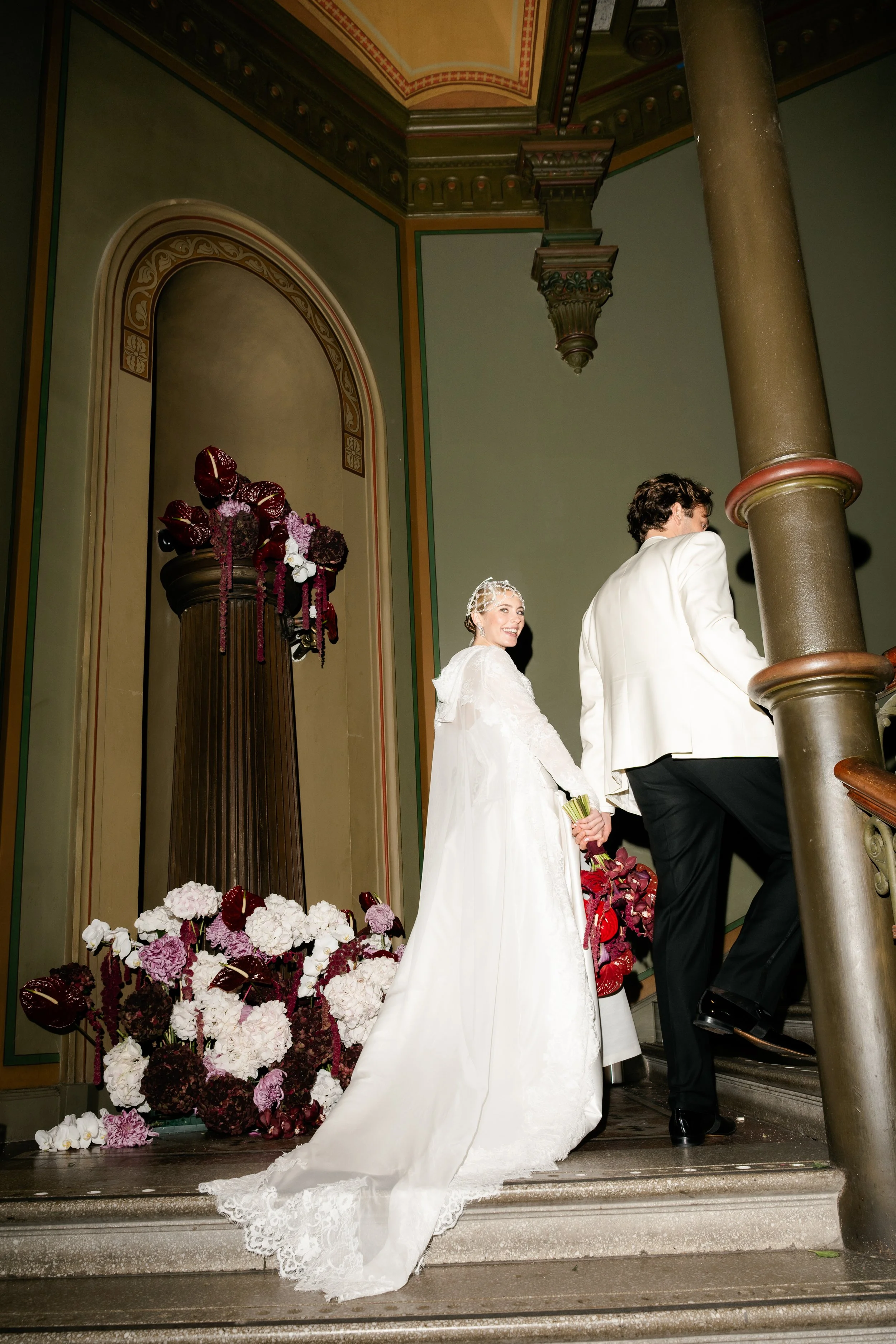 A bride and groom holding hands while ascending a staircase decorated with pink, red, and white flowers, in an elegant interior setting.