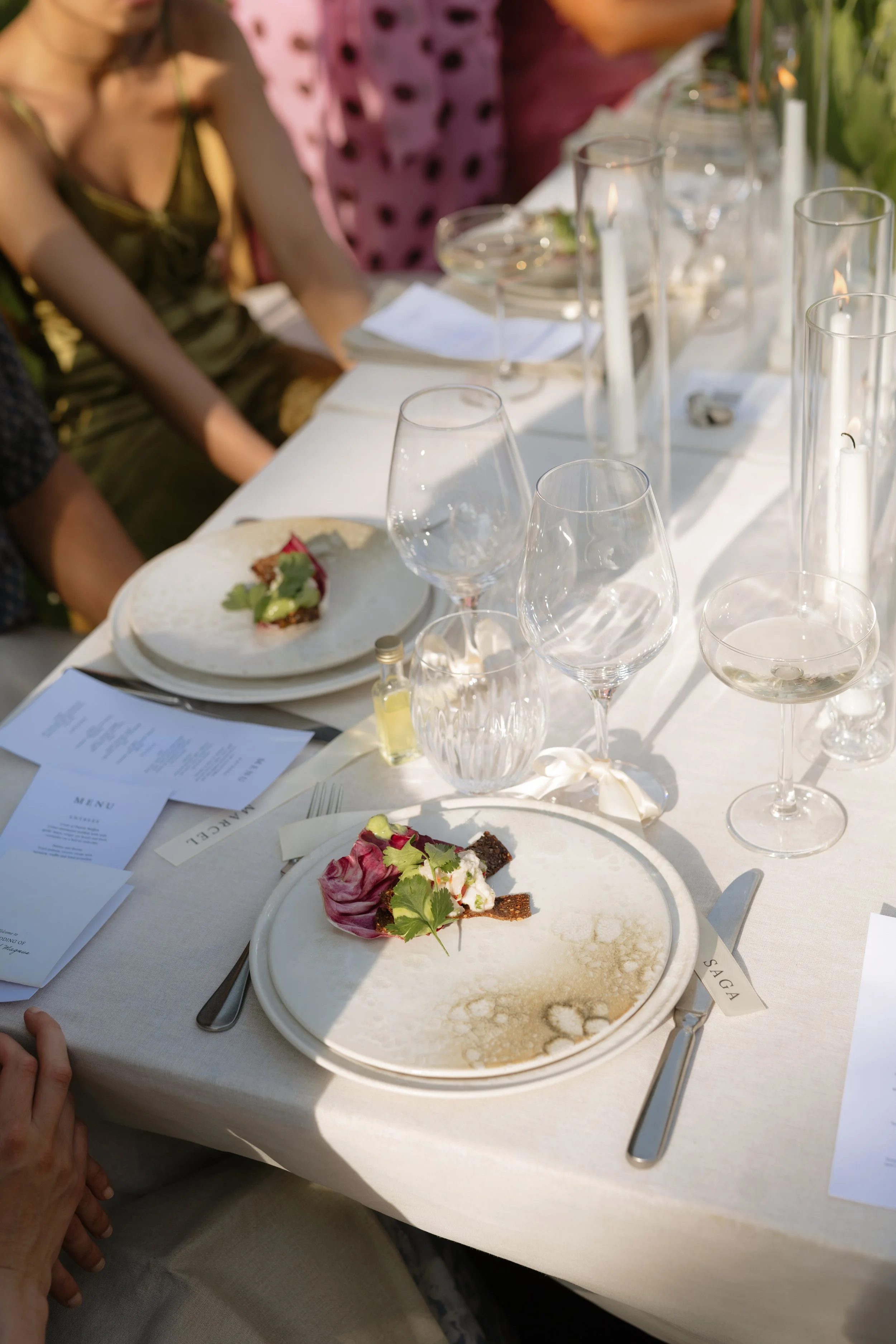 A table set for a meal with empty dessert plates, glasses, and silverware. There are menu cards, candles, and a small bottle of what might be flavored oil or vinegar on the table. The plates have remnants of dessert, decorated with edible flowers and