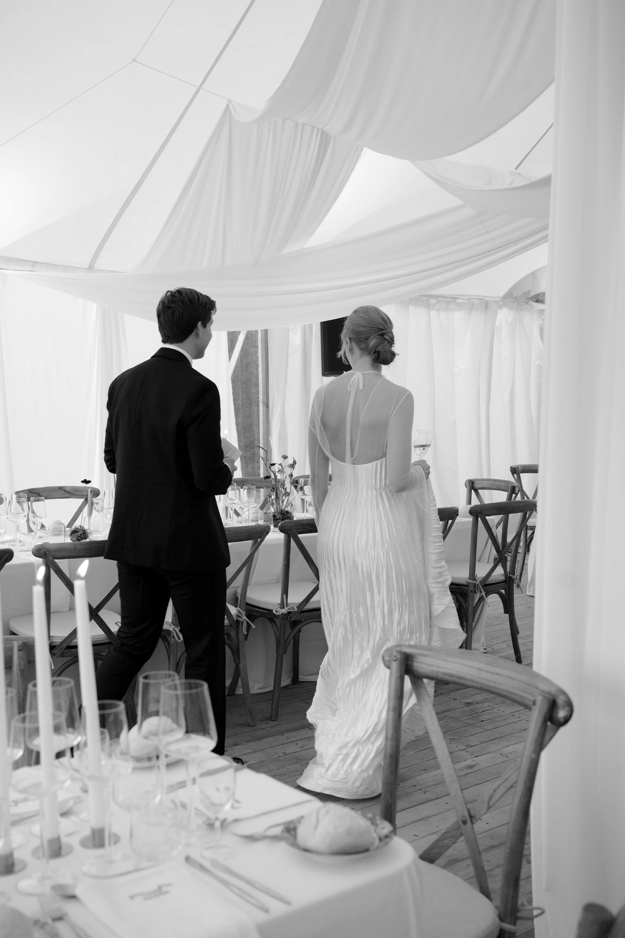 A bride and groom standing in a decorated wedding reception tent, the bride in a white gown holding a glass of wine, and the groom in a black suit holding a small object, with tables set with glasses, candles, and napkins in the foreground.