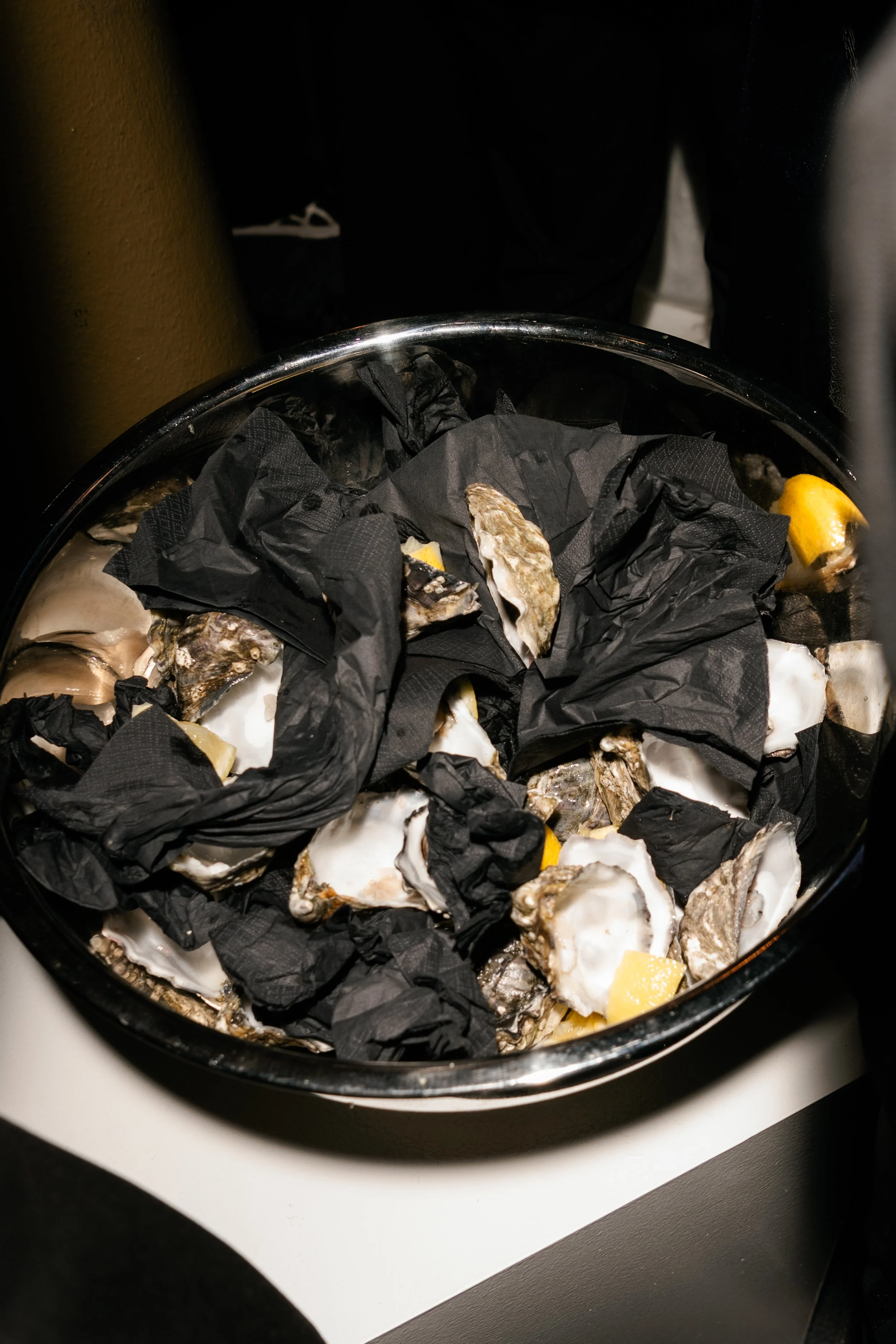 A metal bowl with oyster shells, black tissue paper, and lemon wedges, placed on a white surface.