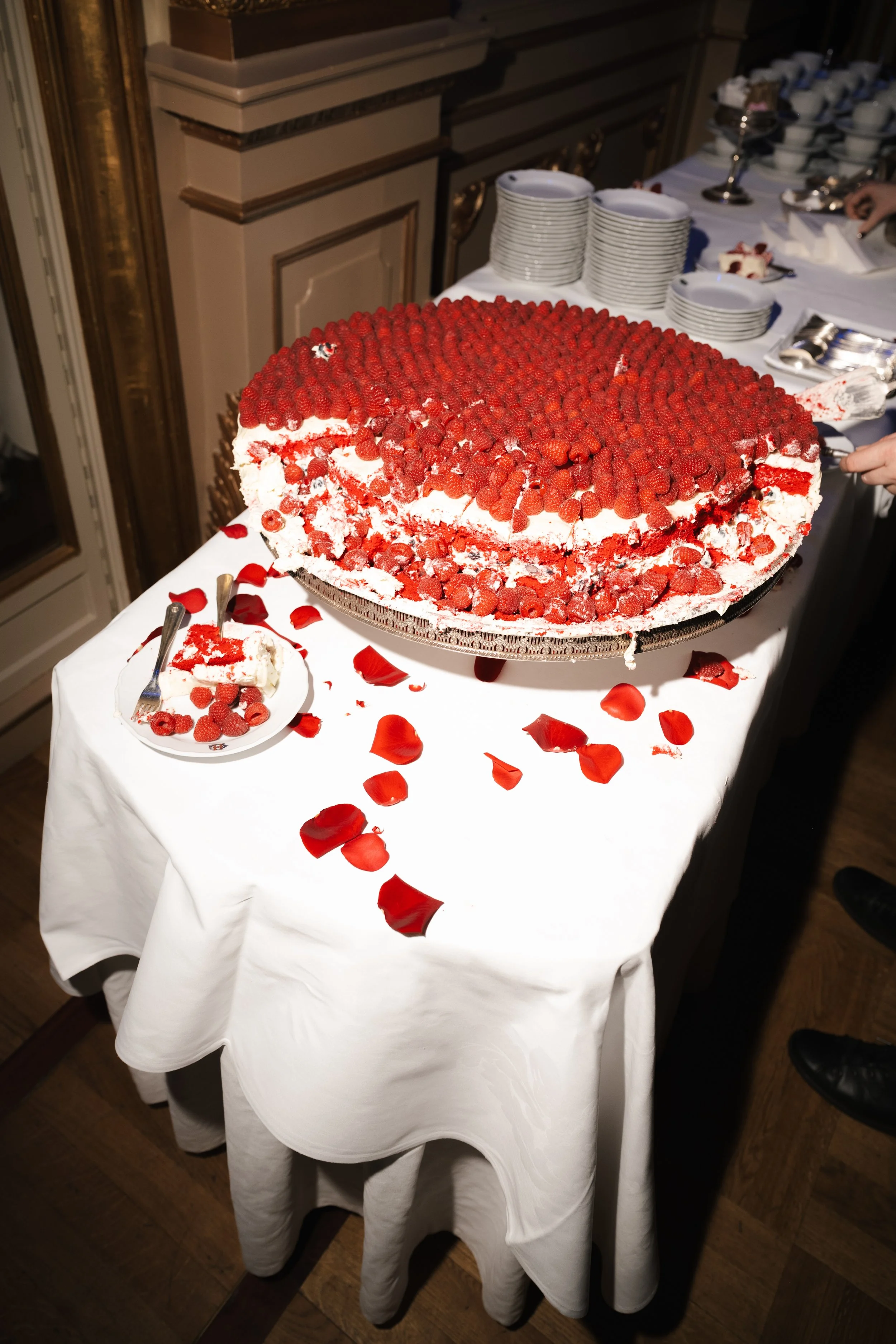 A large strawberry cake on a white tablecloth, topped with fresh strawberries and raspberry decorations, surrounded by scattered strawberry petals and a small plate with a slice of cake.
