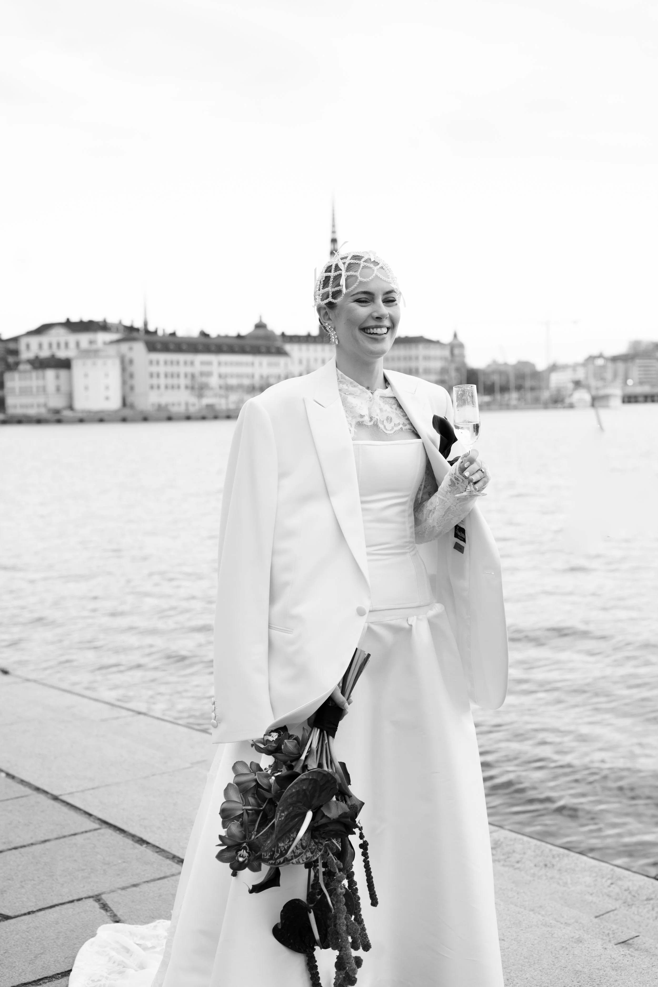 Black and white photo of a smiling woman in a wedding dress, holding a bouquet and a glass of champagne, standing by a waterfront with city buildings in the background.