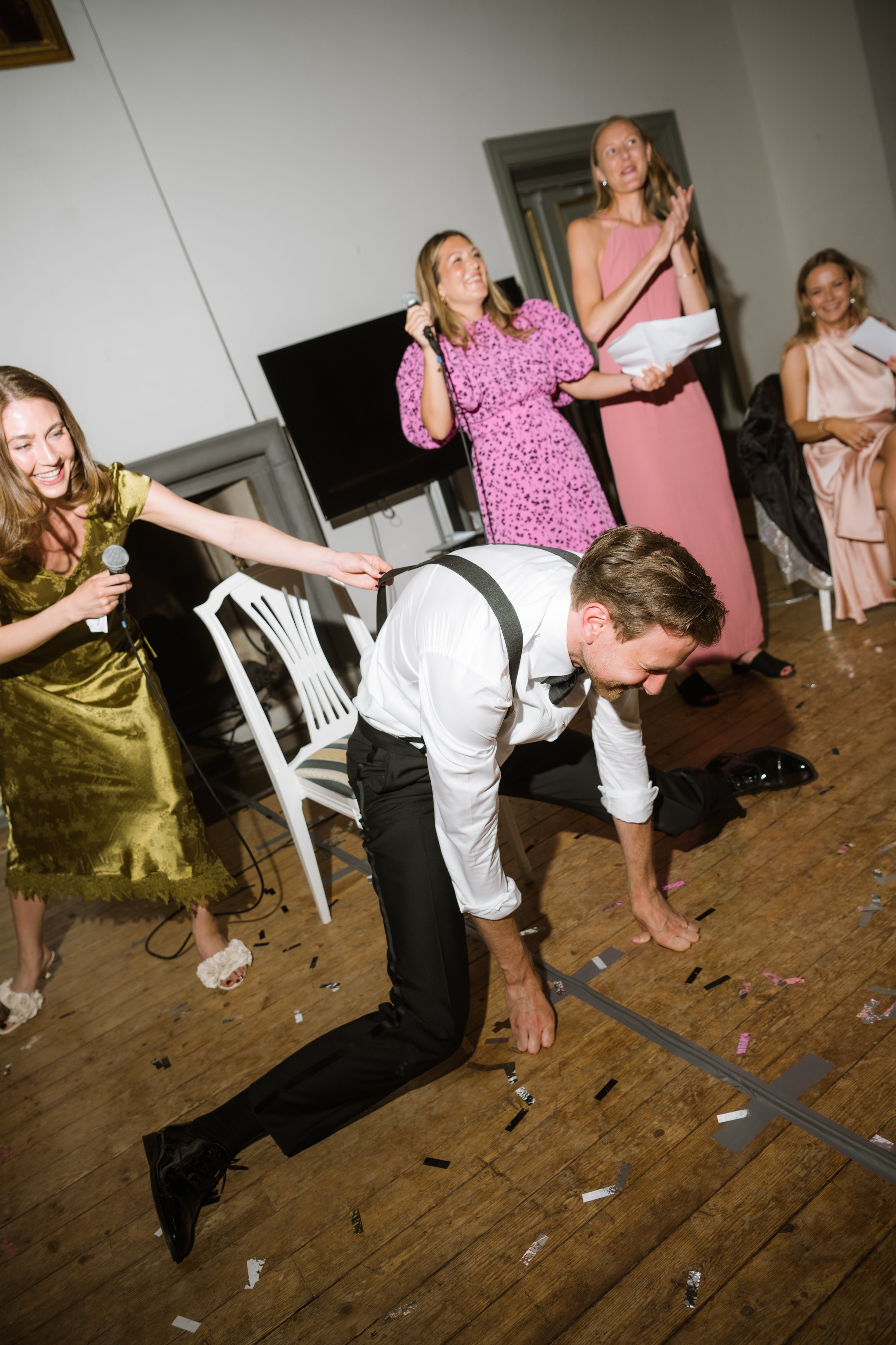A man in formal attire crawling on hands and knees on a wooden floor, surrounded by women in evening dresses, at a party or celebration.