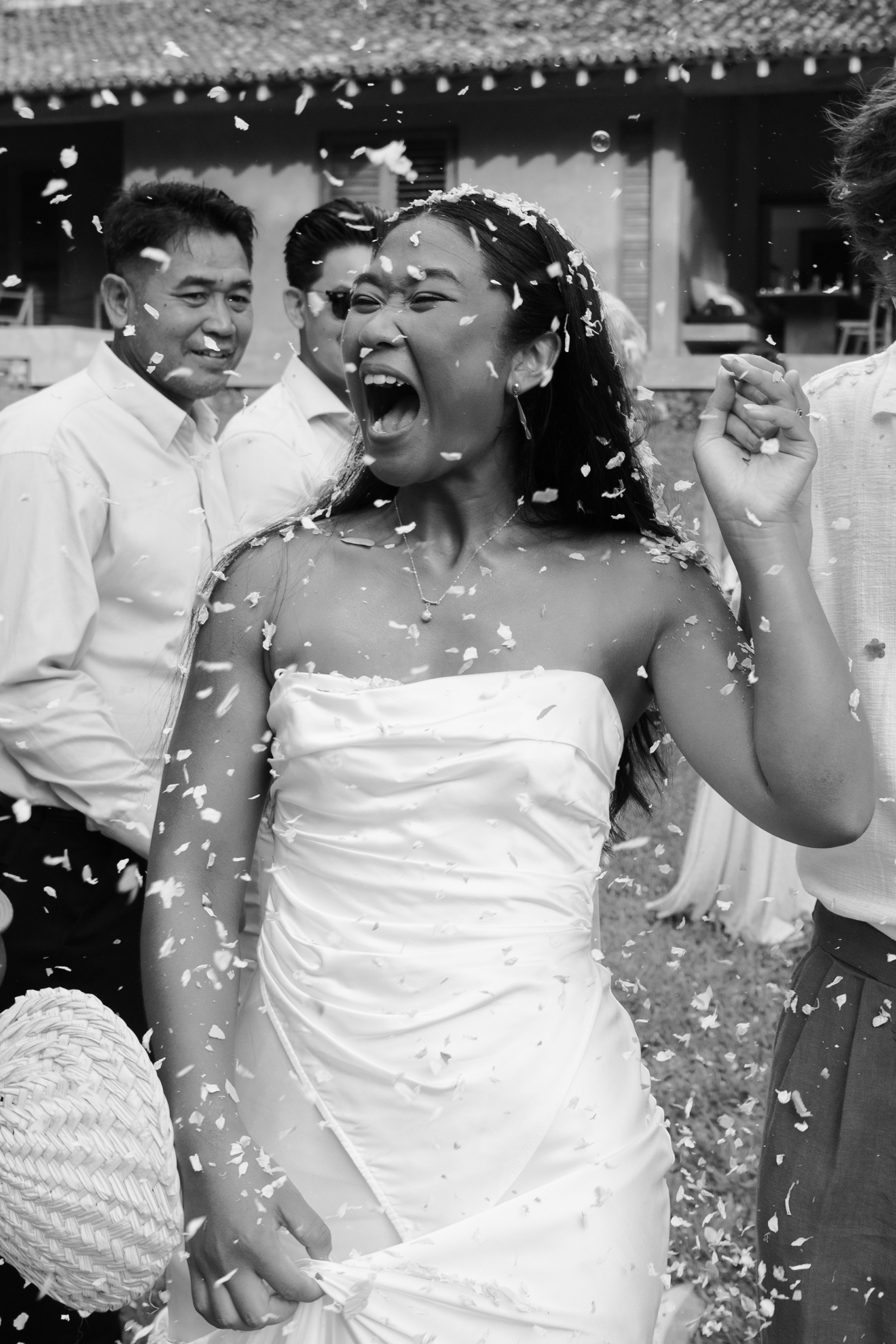 A woman in a wedding dress is celebrating with friends or family at a wedding, surrounded by falling flower petals.