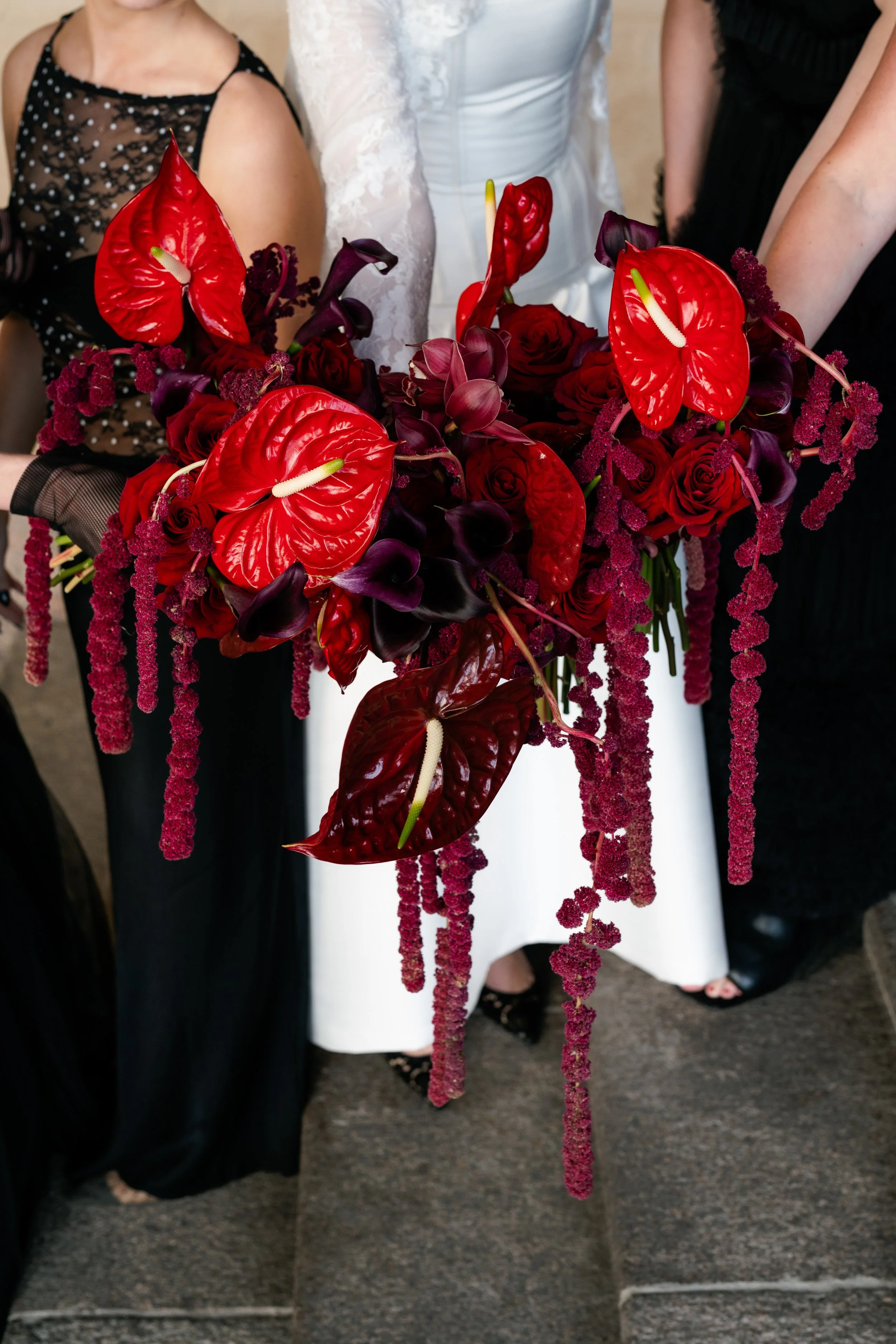 A woman in a white dress holding an elaborate bouquet of red roses, red anthuriums, and purple calla lilies with hanging amaranthus flowers, at a formal event or wedding.