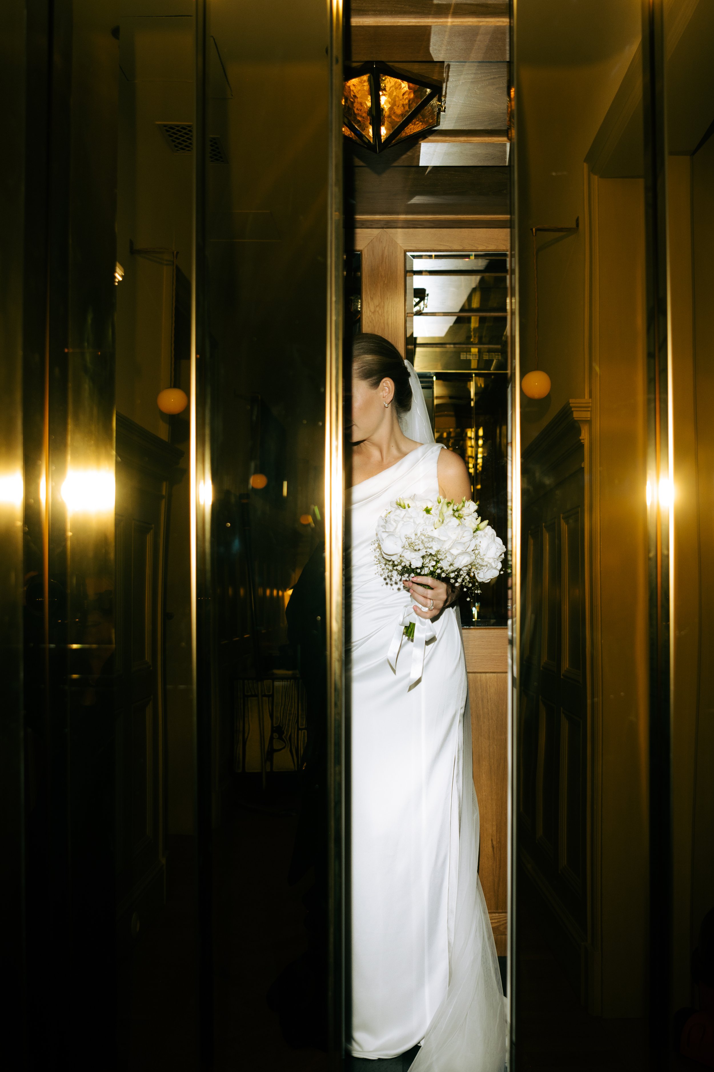 A bride in a white wedding gown holding a bouquet of white flowers, standing in an elevator, partially visible reflected in the mirror.