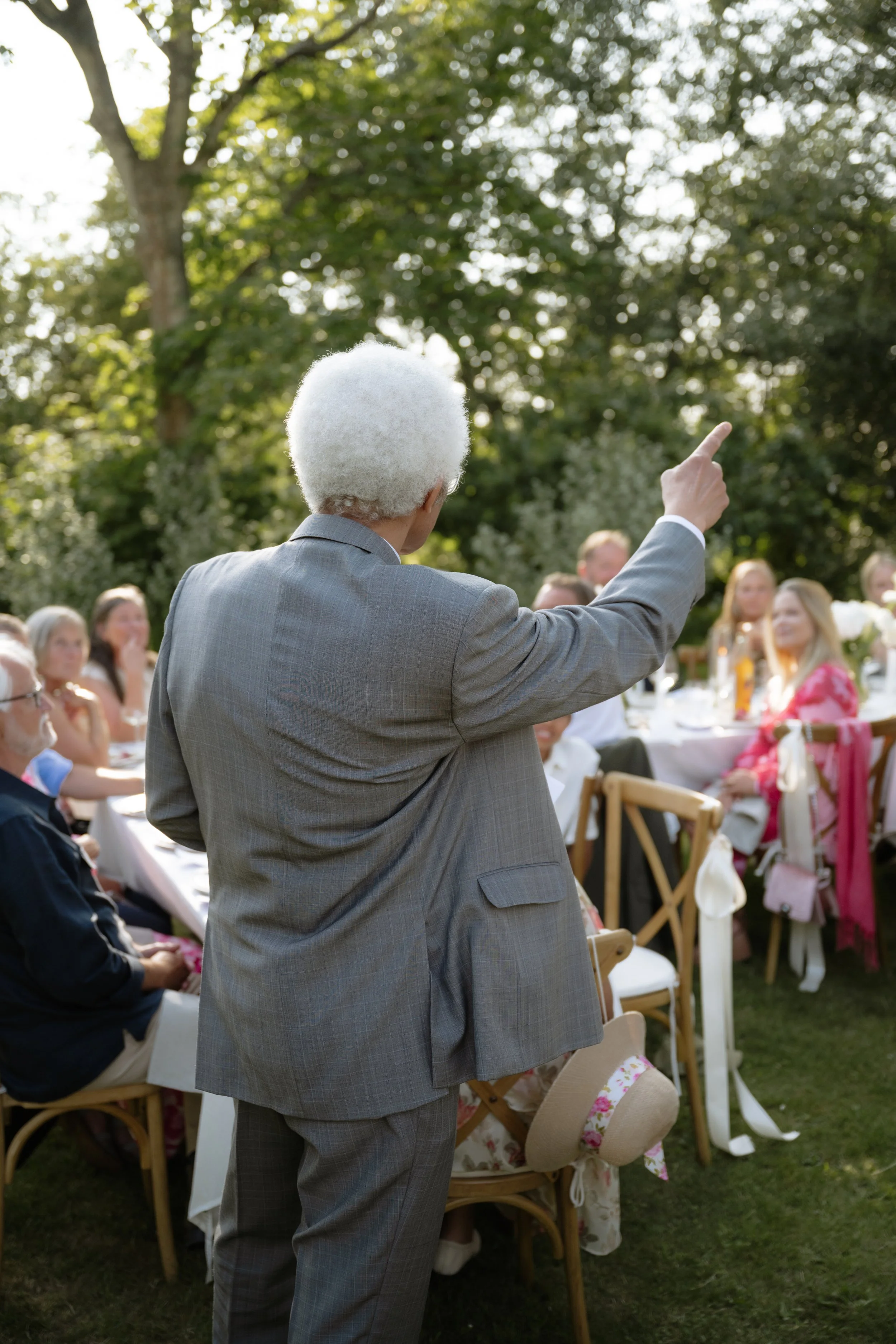 An elderly person with white hair and a gray suit giving a speech at an outdoor gathering with people seated at tables, surrounded by trees.