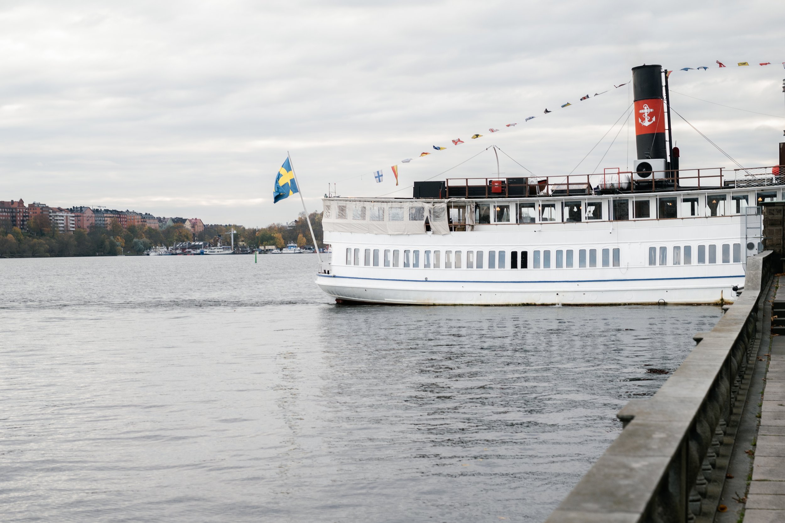 A vintage white boat with a black smokestack and red accents, docked near a waterfront with a cityscape of colorful buildings in the background.