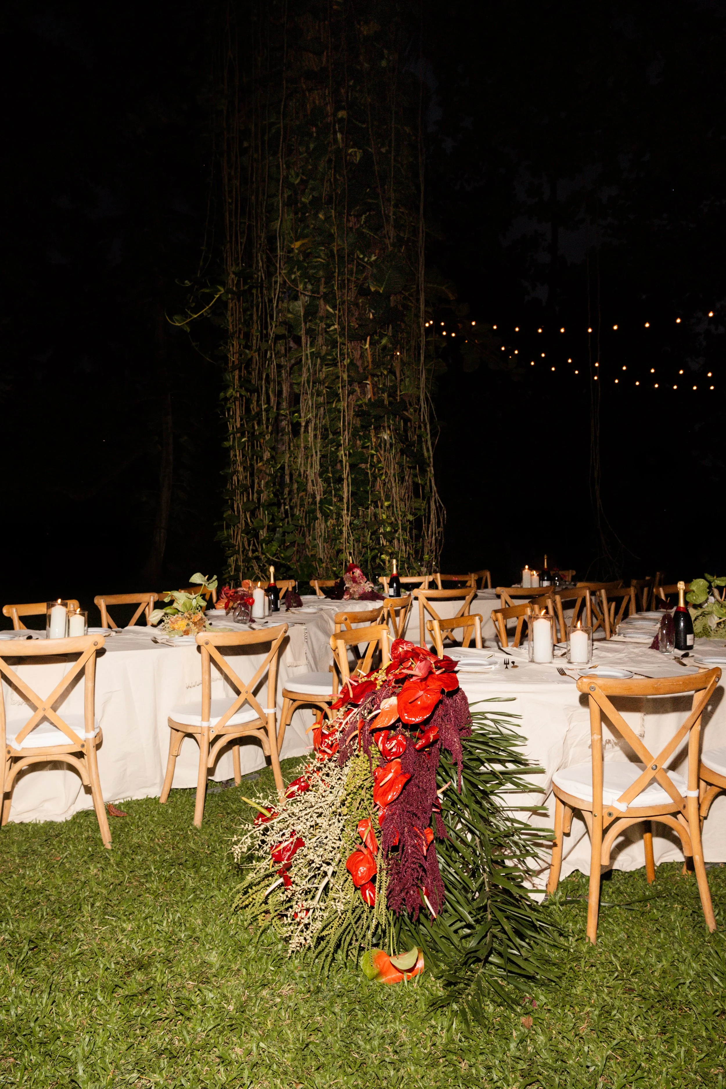 A nighttime outdoor event setup with a long table decorated with floral arrangements featuring red and purple flowers, surrounded by wooden chairs, candles, and small bottles of wine, in a garden setting with large trees and string lights overhead.