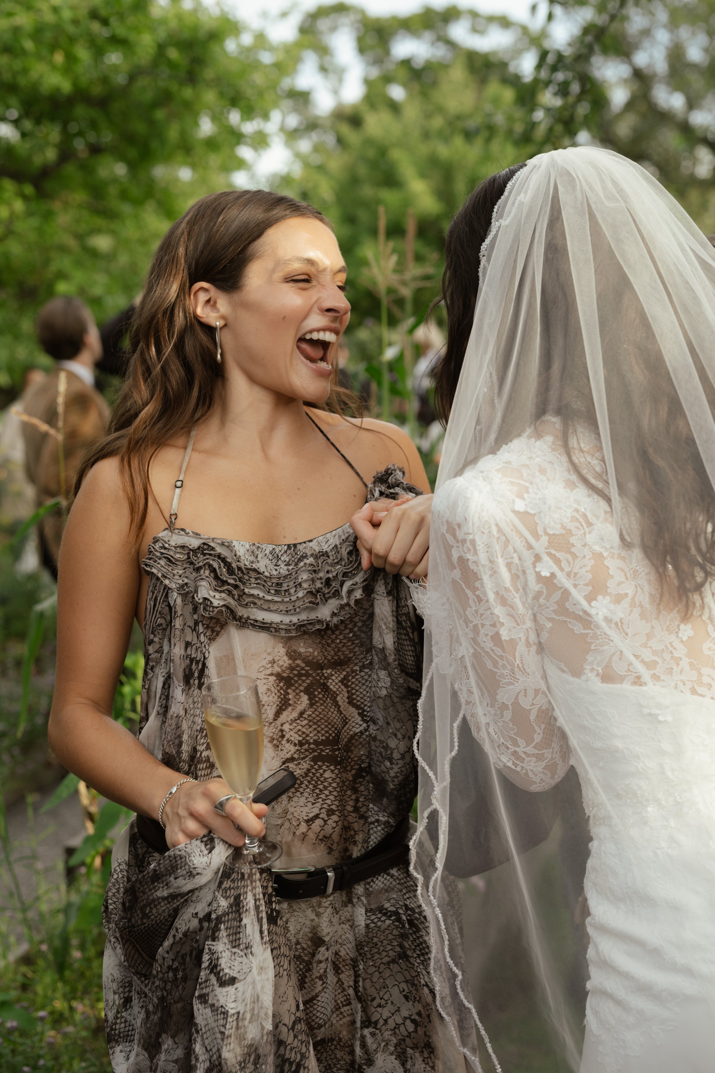 A woman in a snakeskin dress laughing and holding a glass of champagne, standing outdoors with a bride in a lace wedding gown and veil.