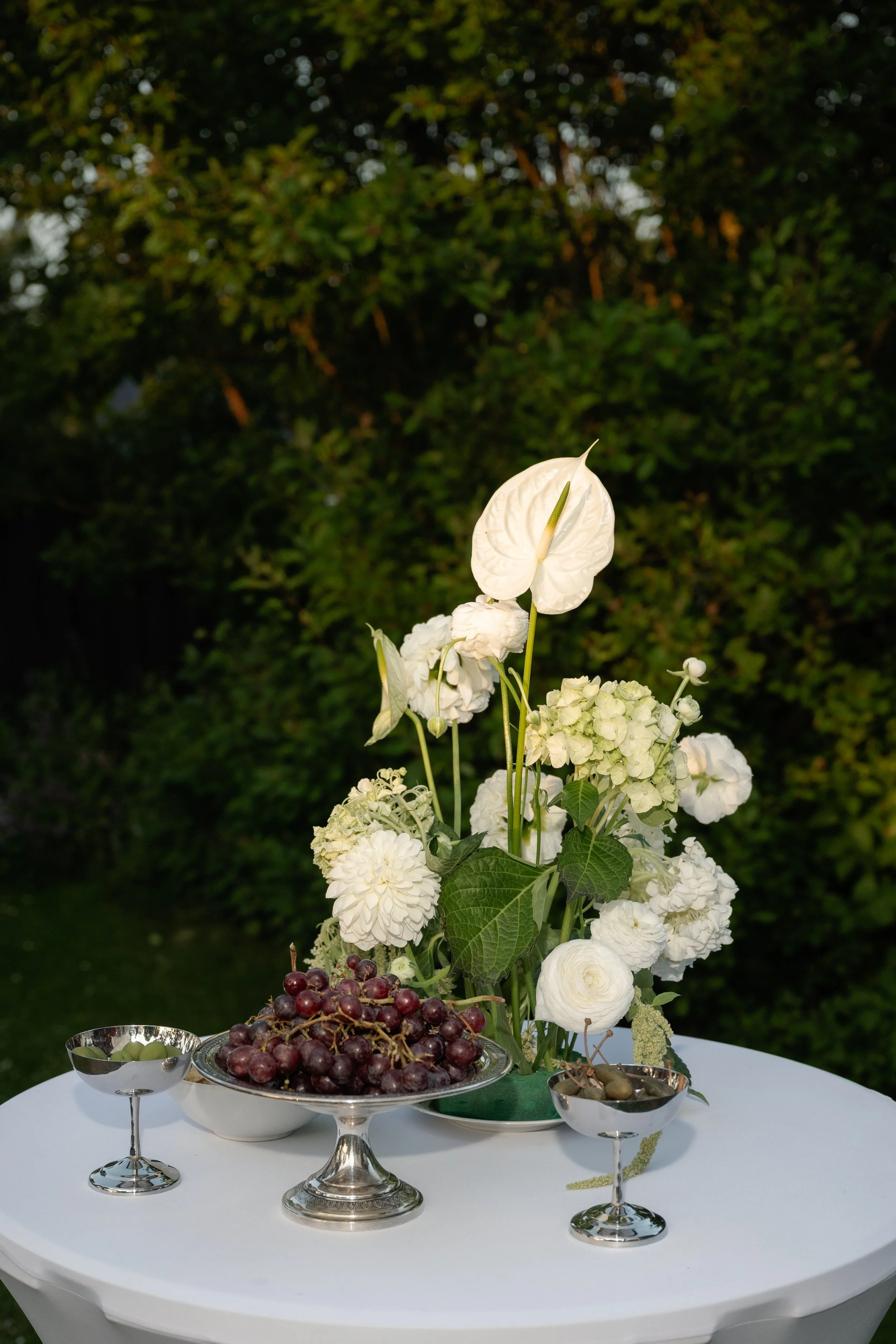 A white table holds a silver dish of red grapes, a small silver bowl of green grapes, and a glass bowl of olives, with a floral centerpiece of white flowers and greenery, set outdoors with green trees in the background.
