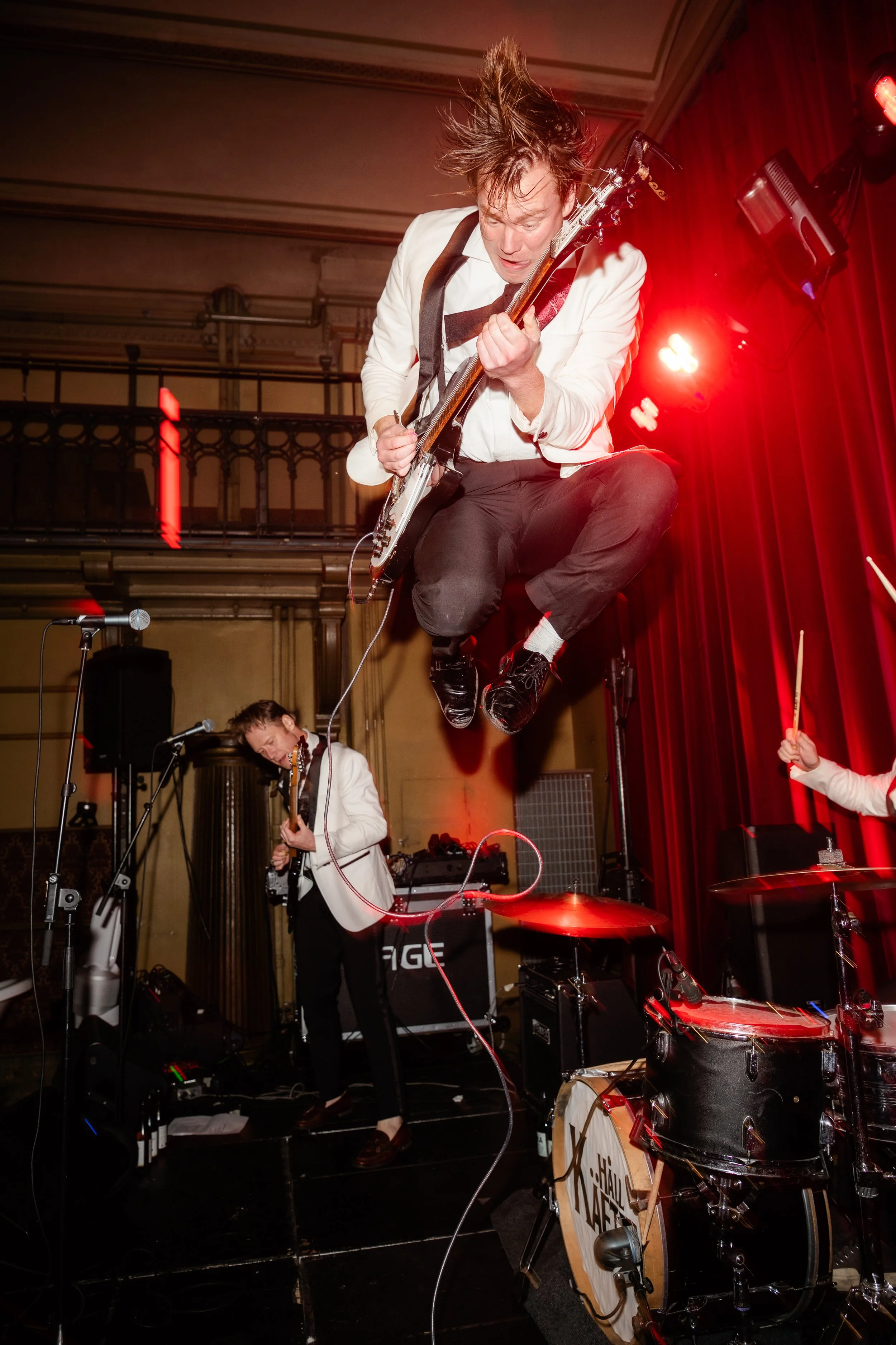 Two musicians performing on stage, one jumping mid-air while playing guitar, and a drummer partially visible.