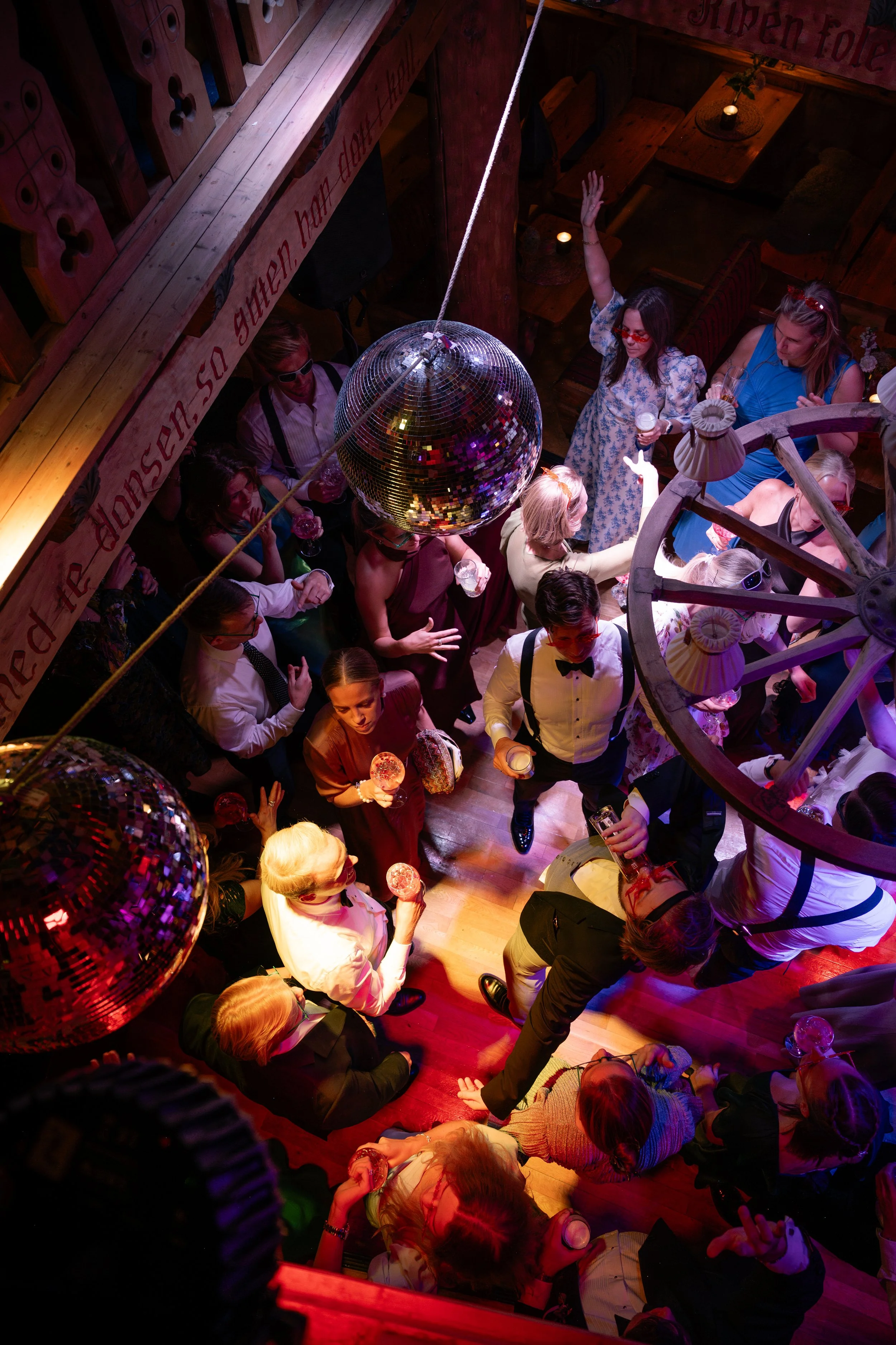 People dancing and socializing at a lively party with disco ball and colorful lighting in a wooden-themed venue.