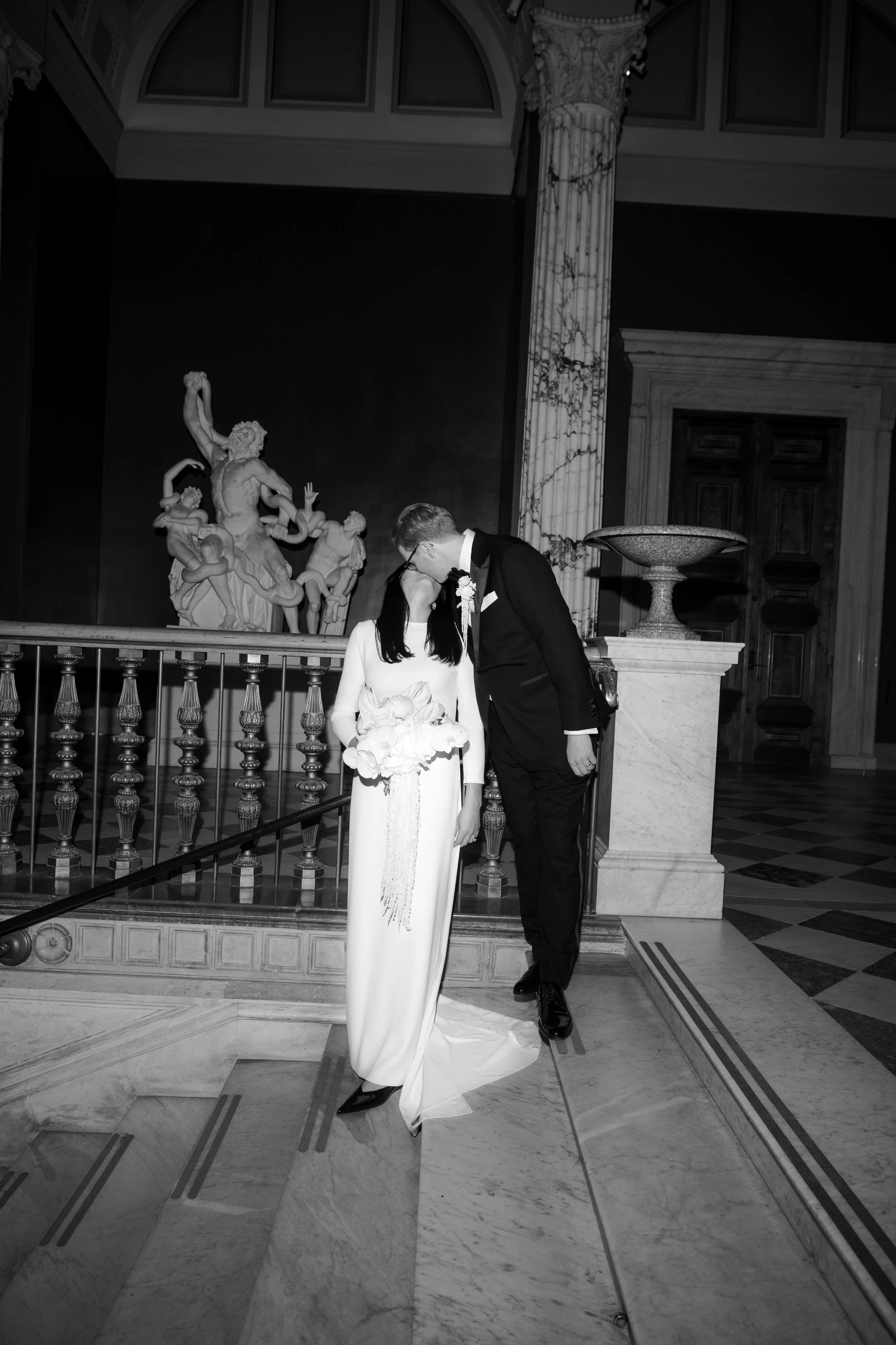 A bride and groom kissing on a staircase inside a historic building, with classical statues and architectural details in the background.