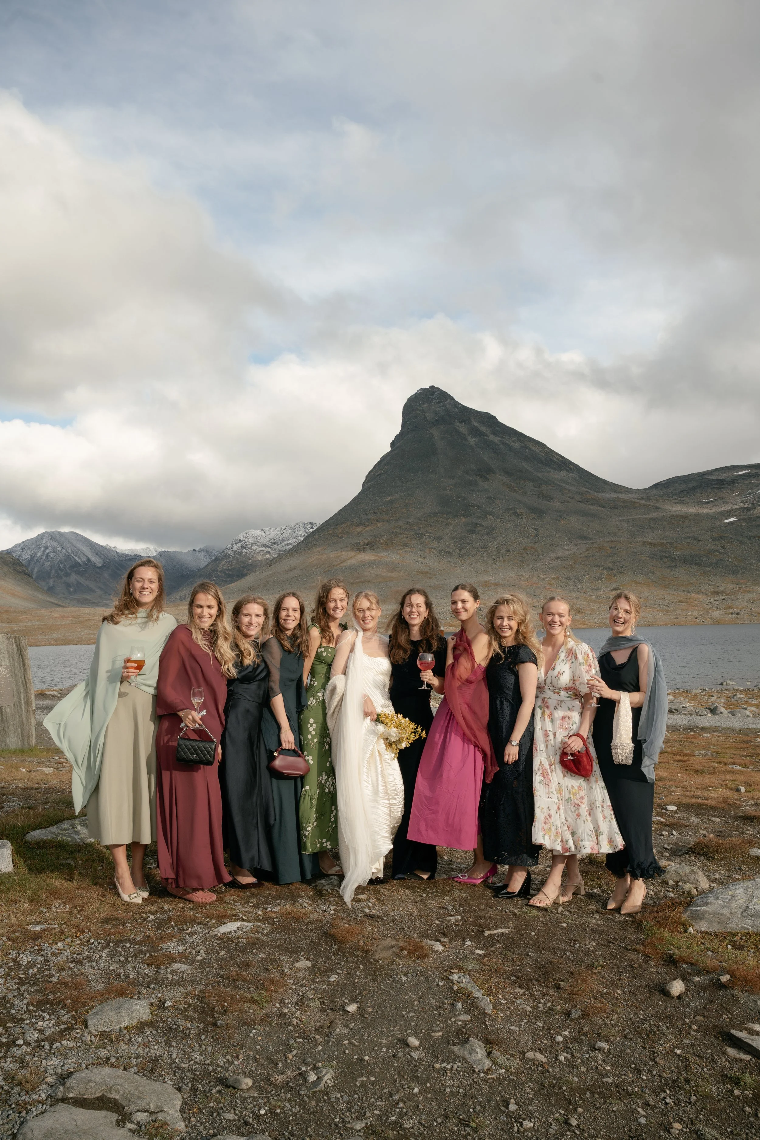 A group of women in elegant dresses standing outdoors near a lake with mountains in the background, celebrating and holding drinks.