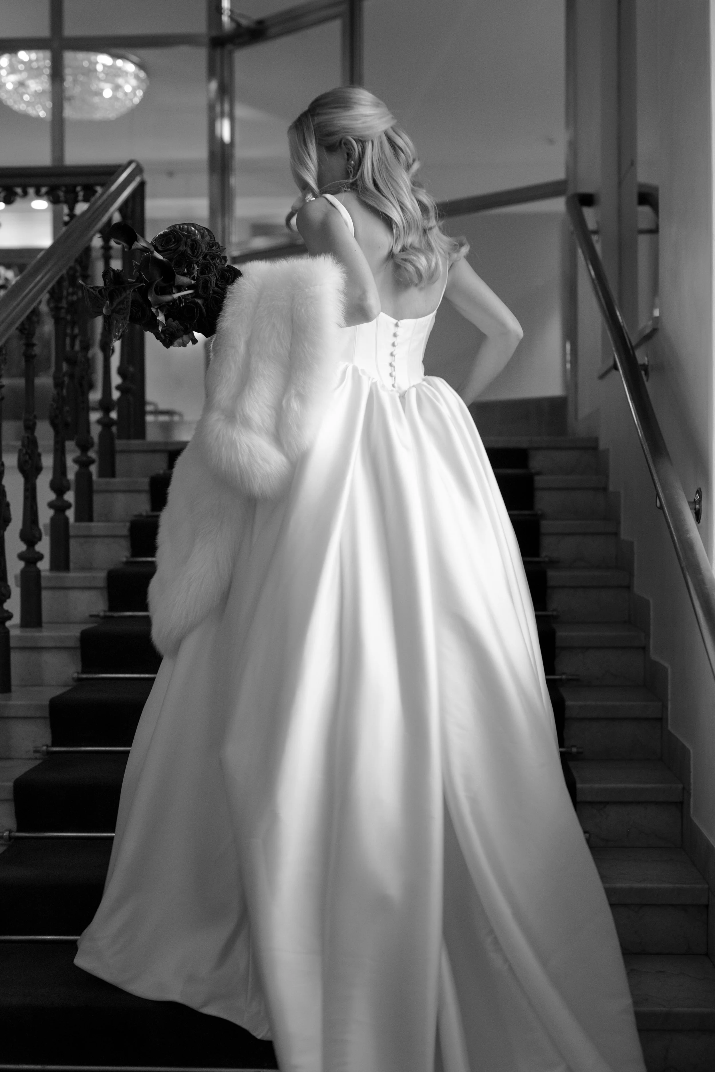 Bride in a strapless wedding gown holding a bouquet of dark roses, descending a staircase indoors.