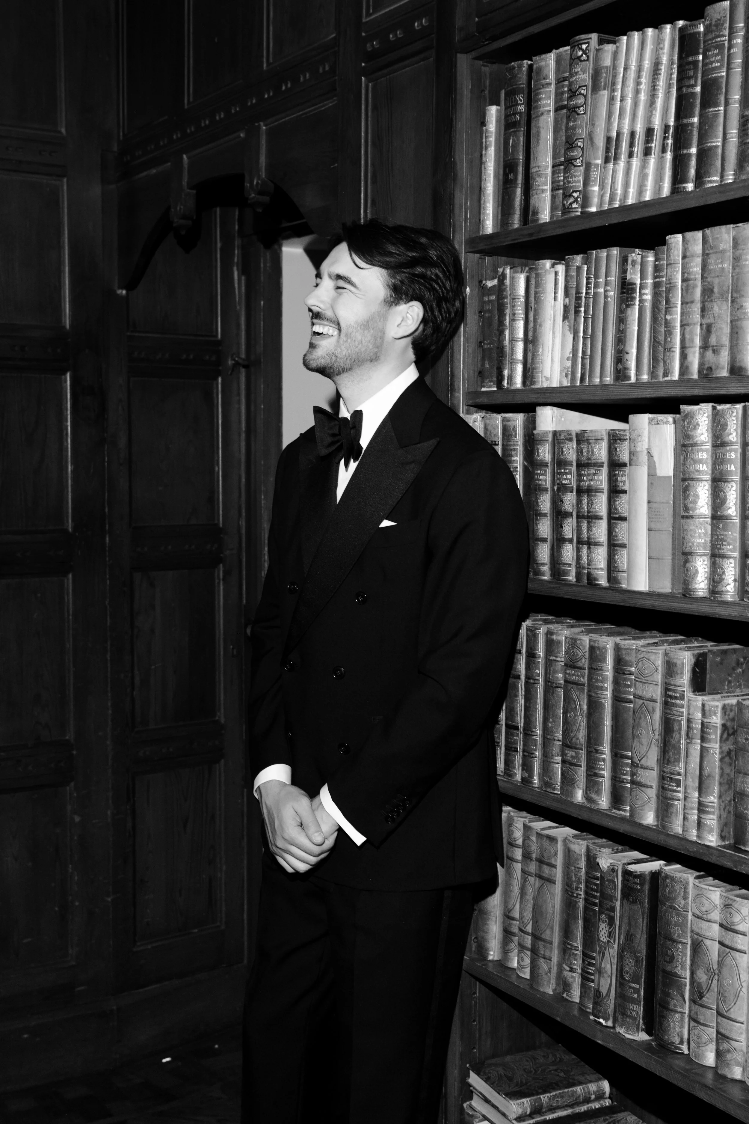 A man in a tuxedo standing in front of bookshelves with leather-bound books, smiling and looking to the left.