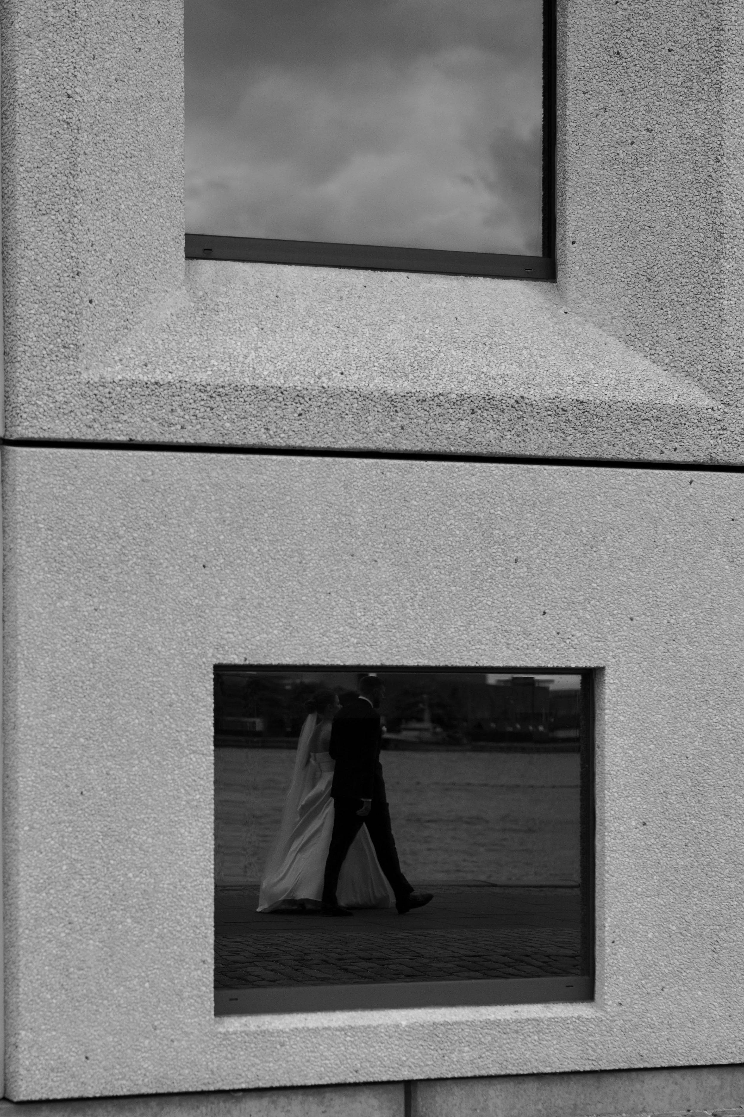 Reflected image of a bride and groom walking near a body of water, seen through two rectangular windows in a concrete building in black and white.