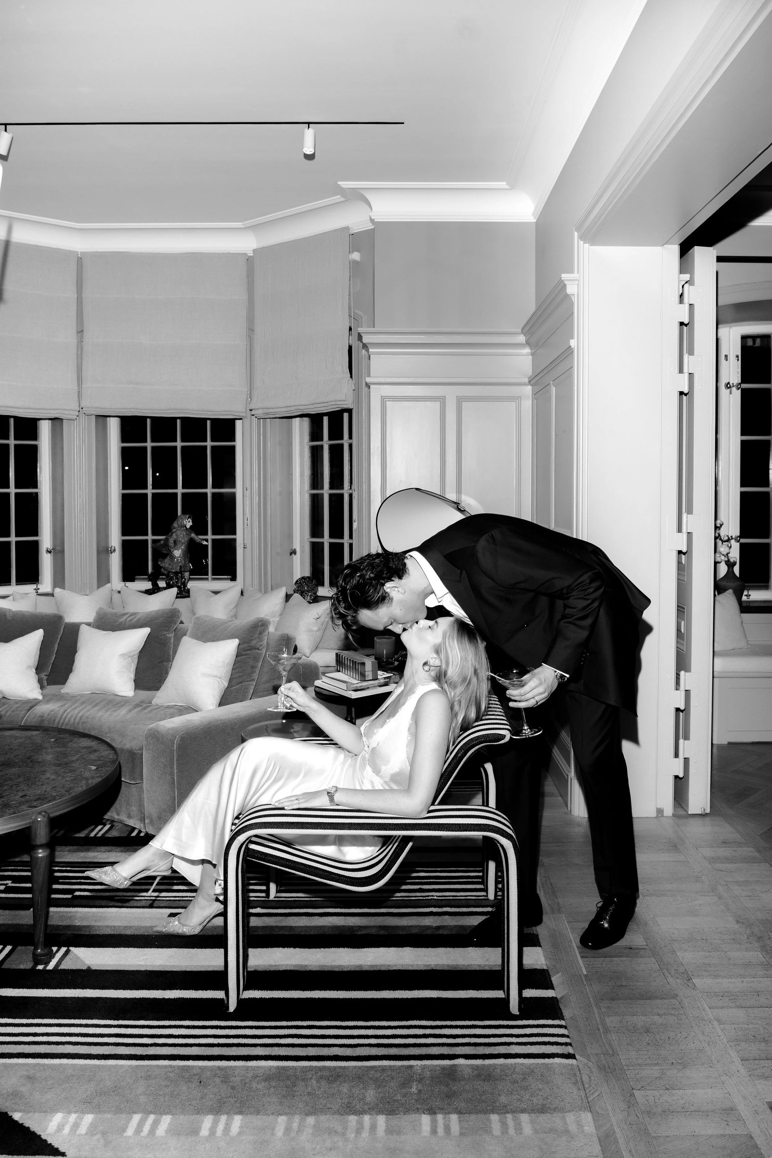 A black and white photo showing a romantic moment between a man in a suit and a woman in a satin dress, with the man leaning over to kiss the woman who is sitting on a striped chair, holding a glass of wine in her hand.