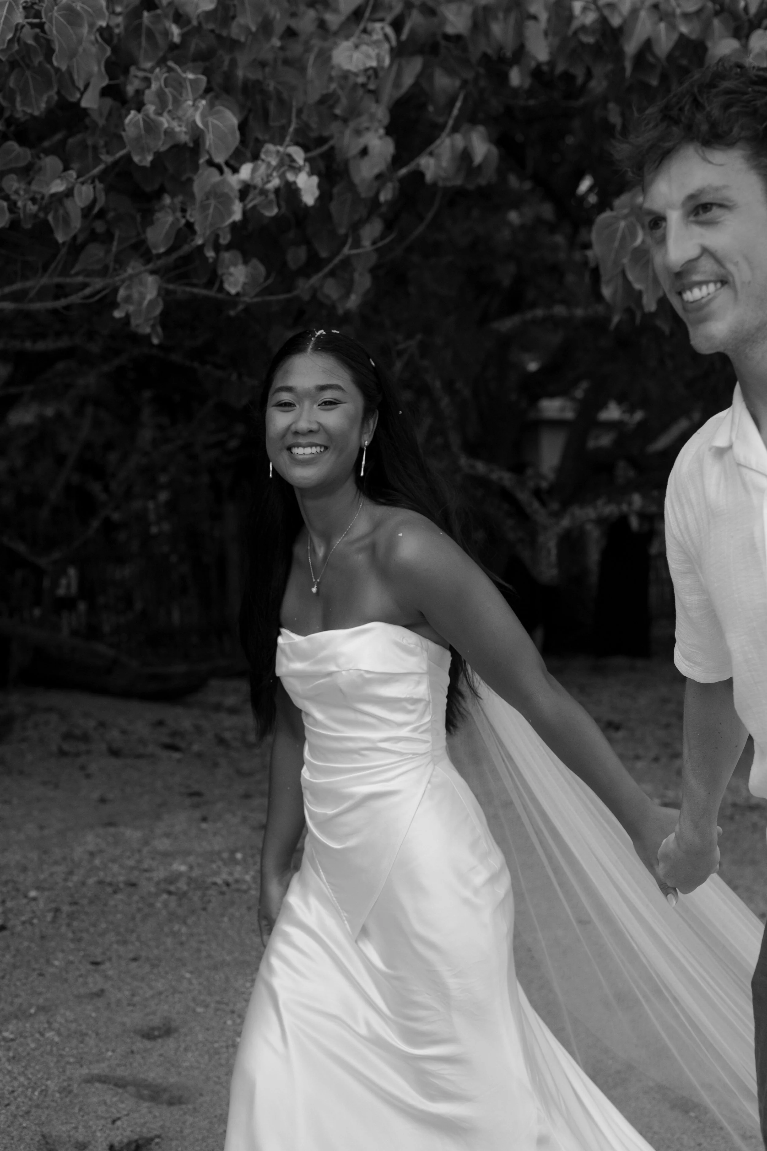 A black-and-white photo of a smiling bride in a strapless wedding gown holding hands with a man, both outdoors with trees in the background.