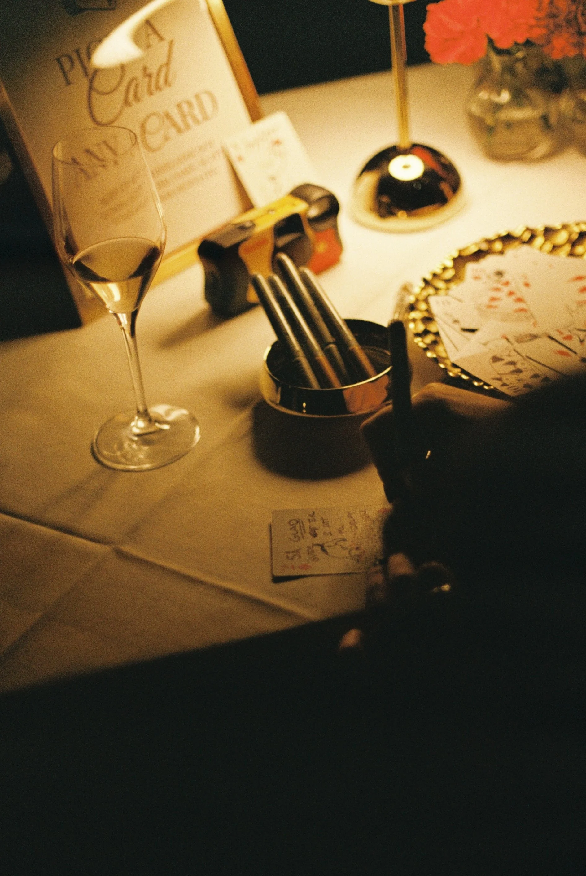 A dimly lit table with a glass of champagne, playing cards, a small note, pens in a container, and a hand holding a pen is visible. There is a framed sign, a lamp, and a flower arrangement in the background.
