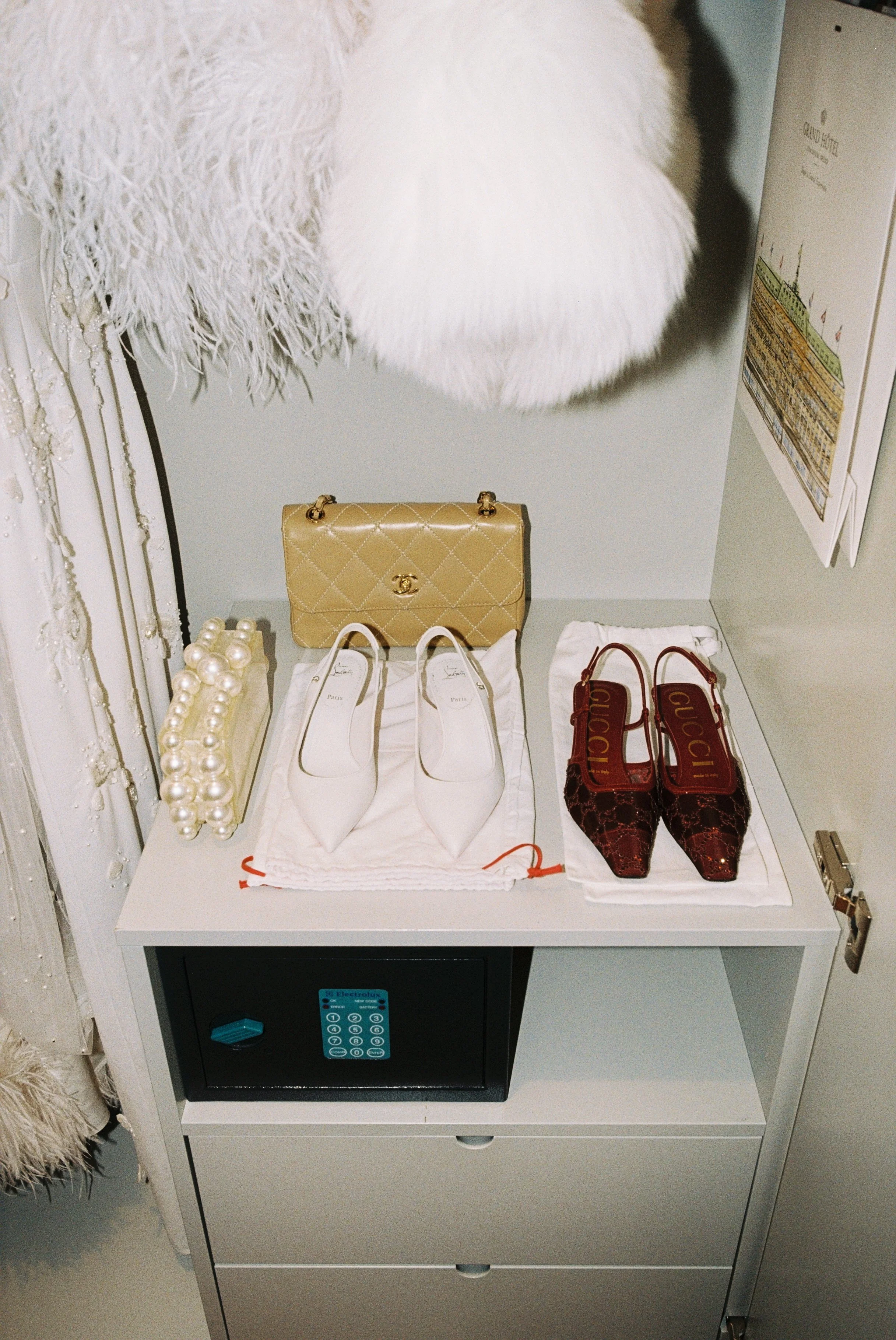 A white cabinet with three drawers holding a black safe, topped with a display of two pairs of designer shoes—white pointed-toe flats and dark red Gucci heels—along with a yellow Chanel handbag and a pearl clutch purse, all on white cloths, with a fu