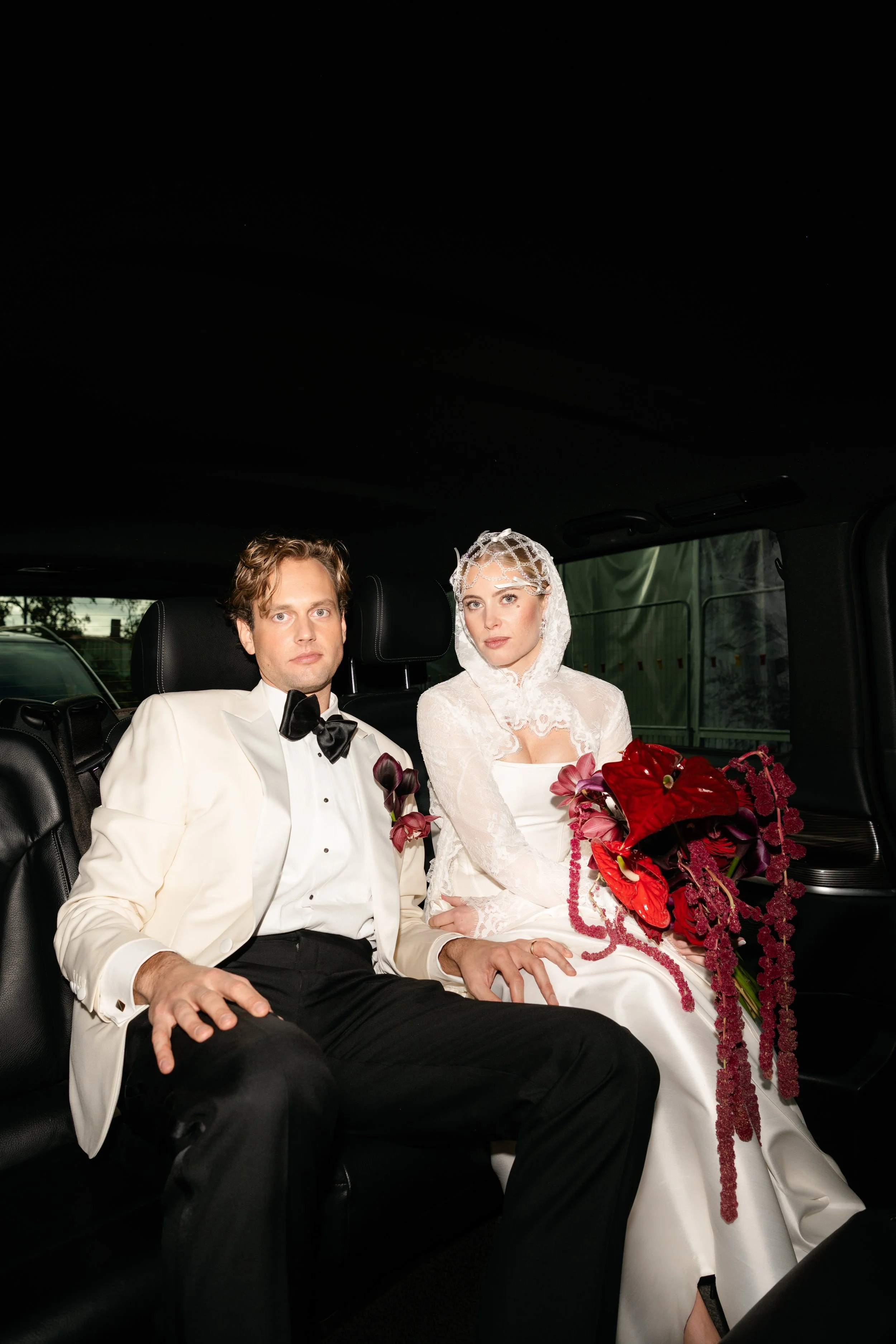 A bride and groom sit inside a vehicle, with the bride holding a large bouquet of red and purple flowers. The groom is wearing a white tuxedo jacket, black pants, and a black bow tie, while the bride is in a lace white dress with a lace veil.