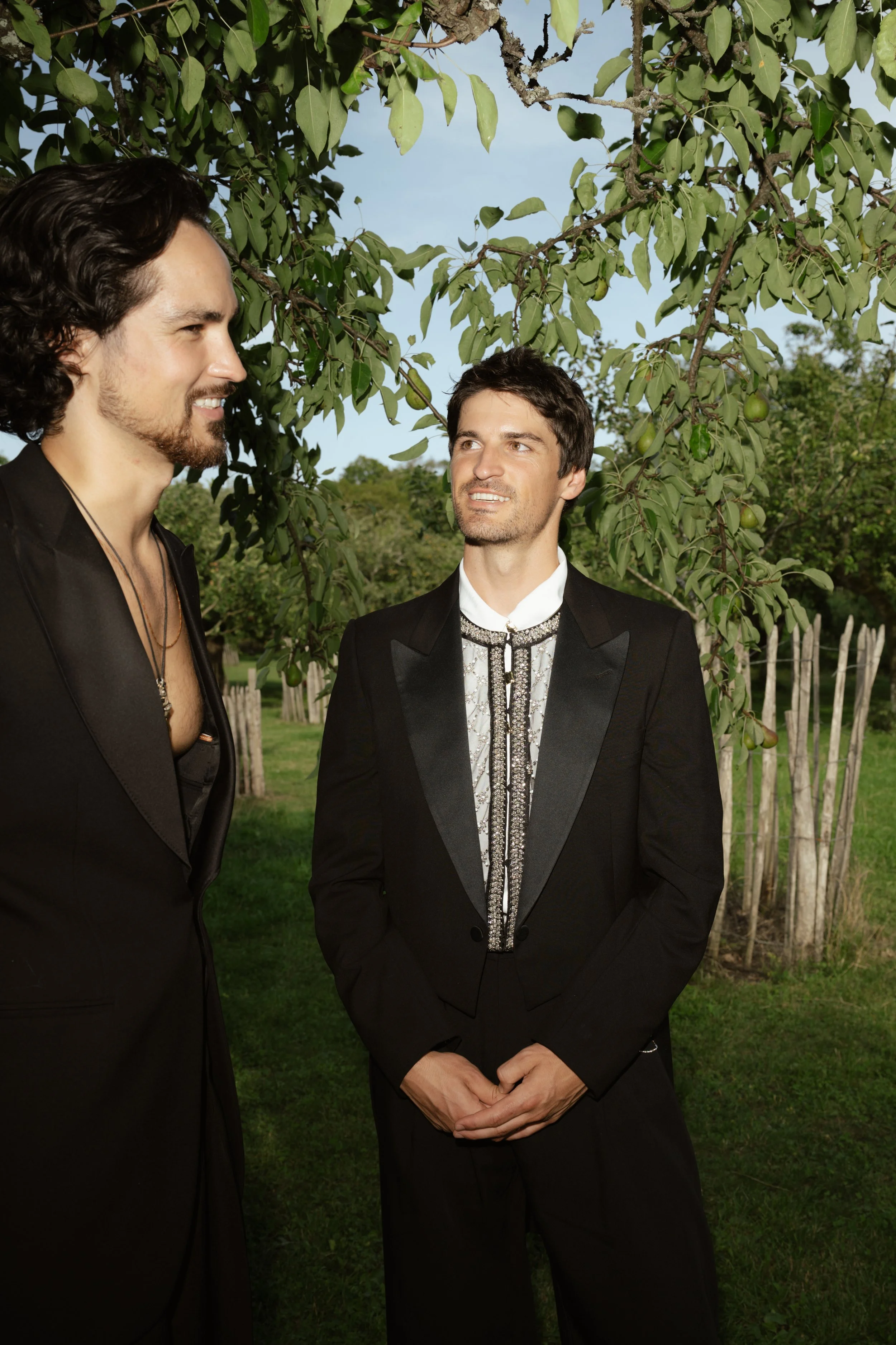 Two men dressed in formal black suits standing outdoors under a tree, engaged in conversation. One man has long, wavy dark hair and a beard, while the other has short dark hair and no beard.