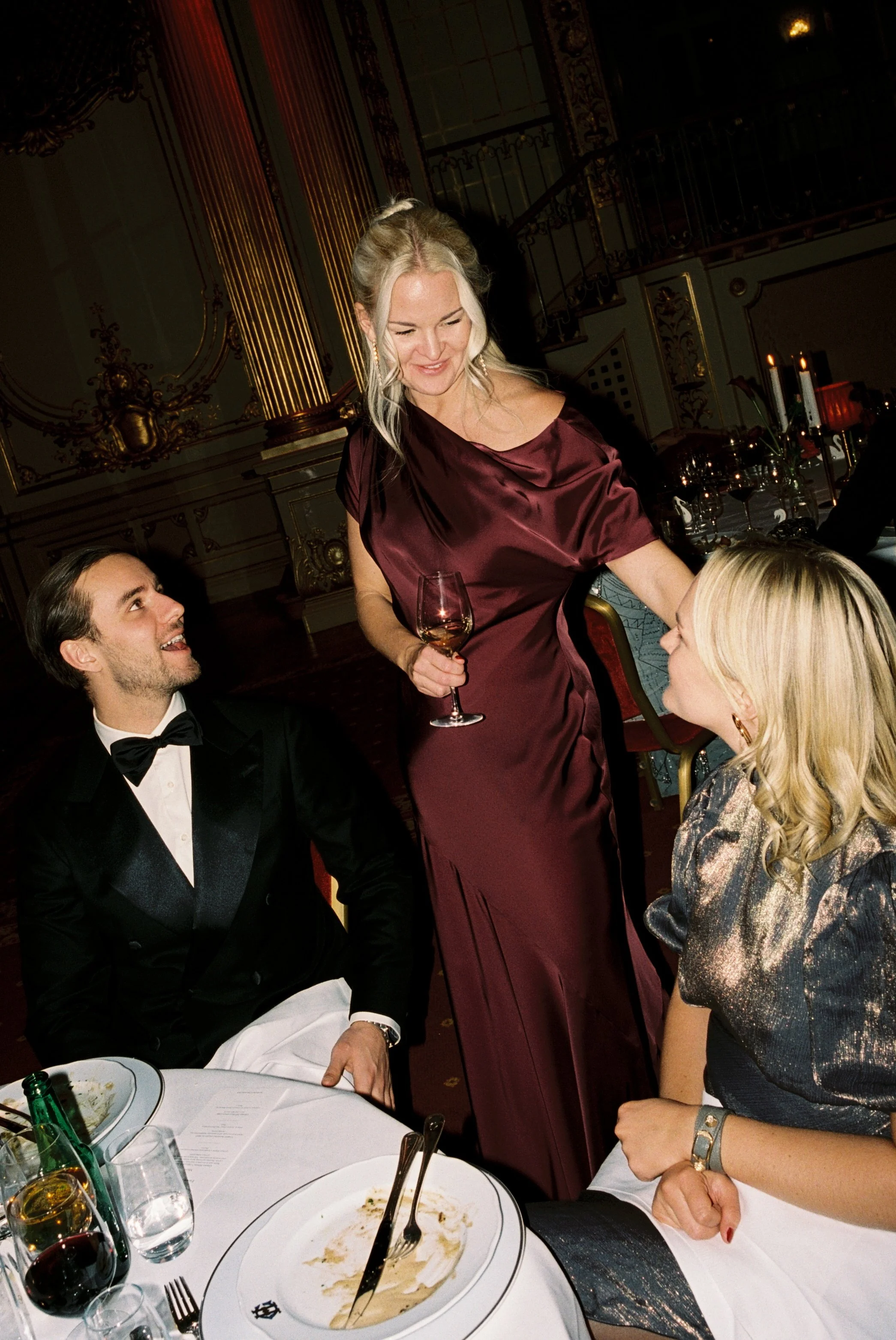 Group of three people sitting at a dinner table, with a woman standing and smiling while holding a glass of wine, engaging with a seated woman and man in a formal setting with ornate decor in the background.