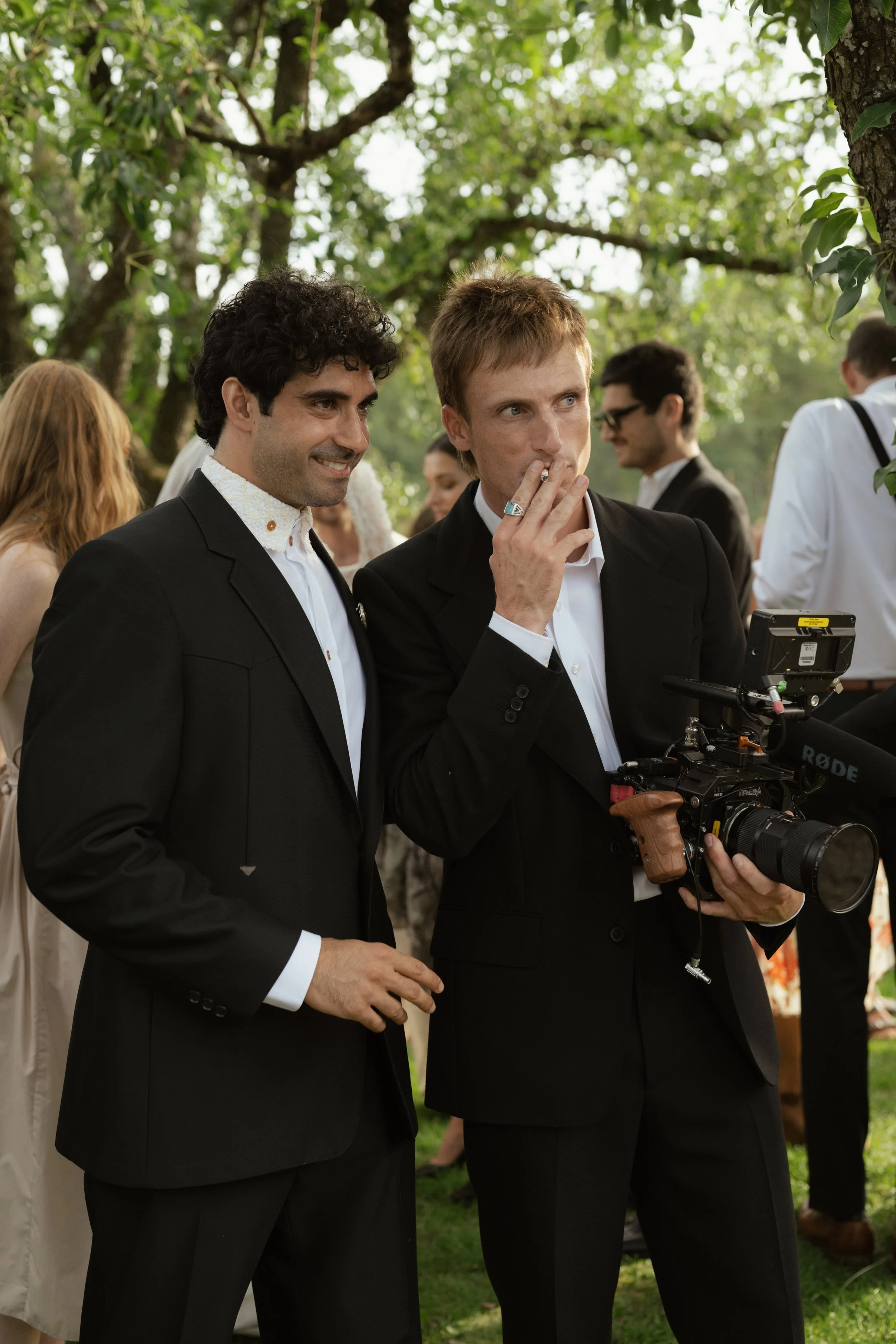 Two men in black suits holding a camera at an outdoor gathering under trees, with other people in the background.