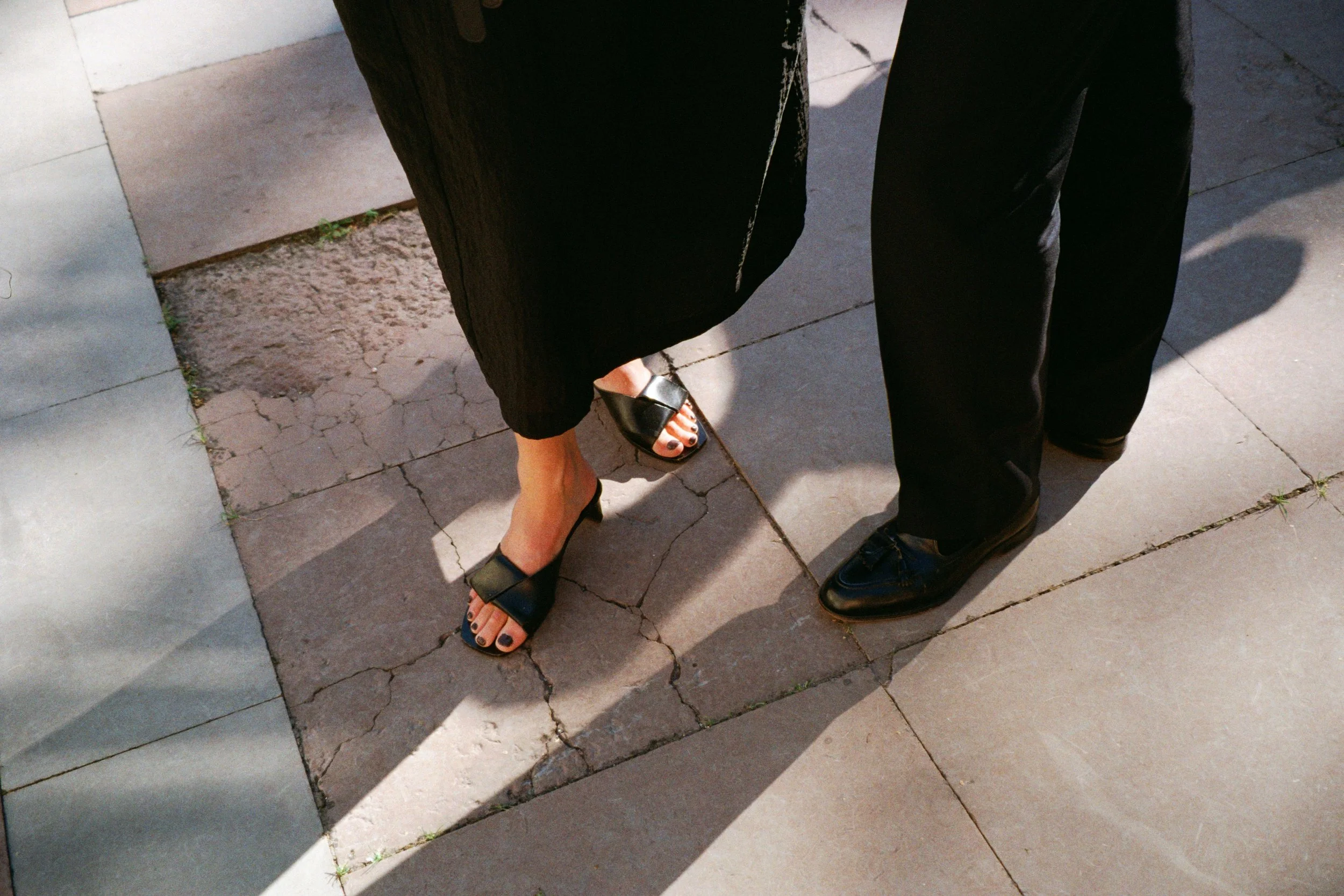 Close-up of two people standing on pavement, showing their legs and feet. One person is wearing black sandals and a black dress, the other is wearing black shoes and black pants.
