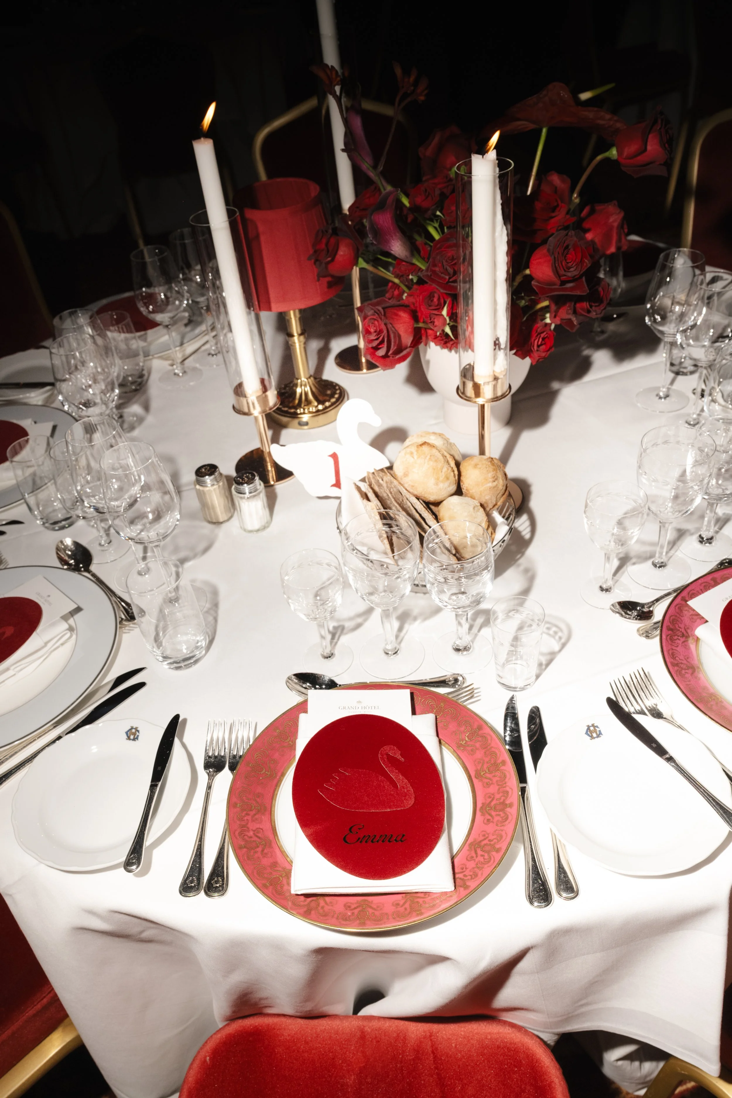 Elegant dining table set for a special occasion with white tablecloth, red and gold accents, red plates, glassware, candles, and a floral centerpiece with red roses and dark purple flowers.