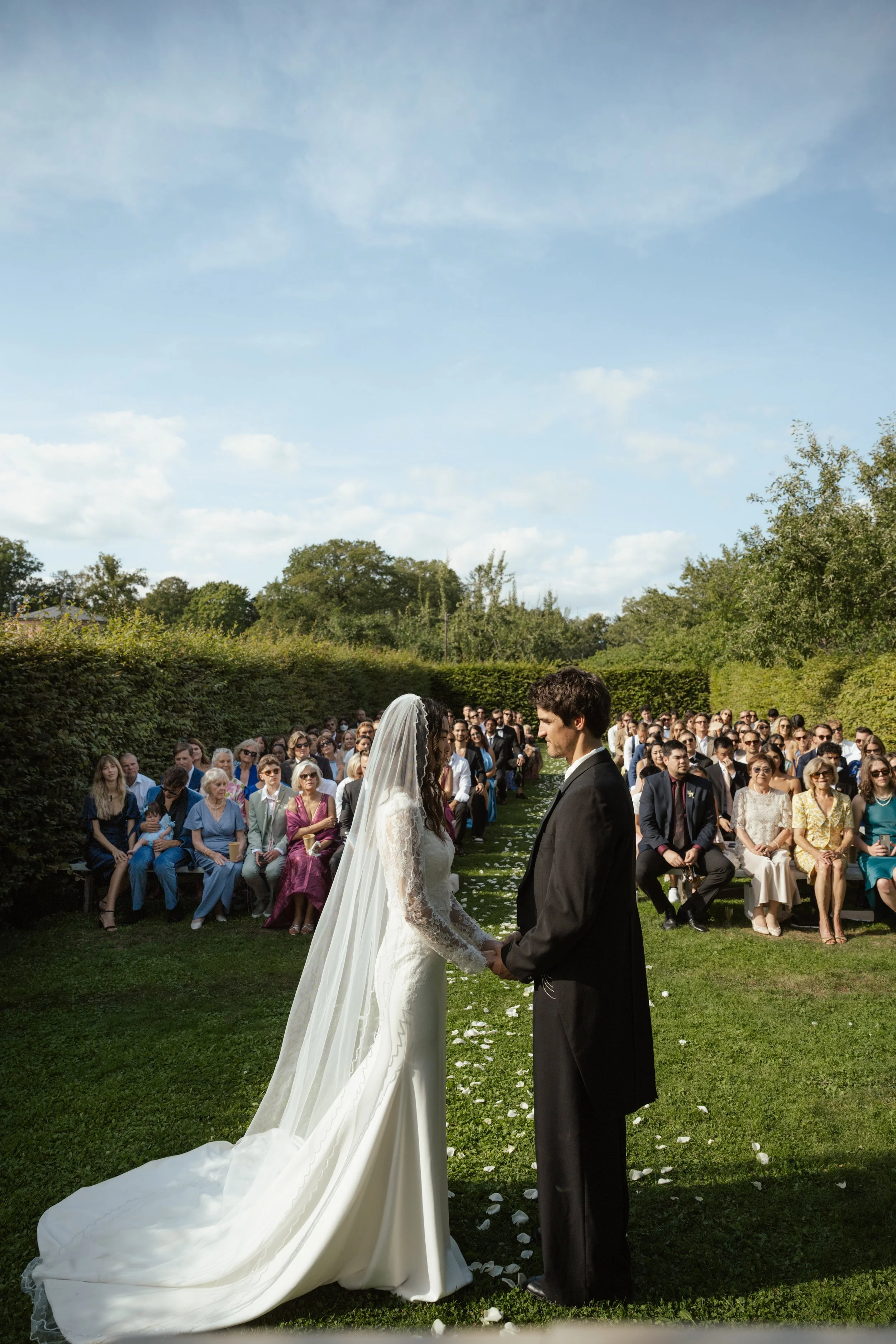 A bride and groom holding hands at an outdoor wedding ceremony, with guests seated in the background on a grassy lawn under a partly cloudy sky.