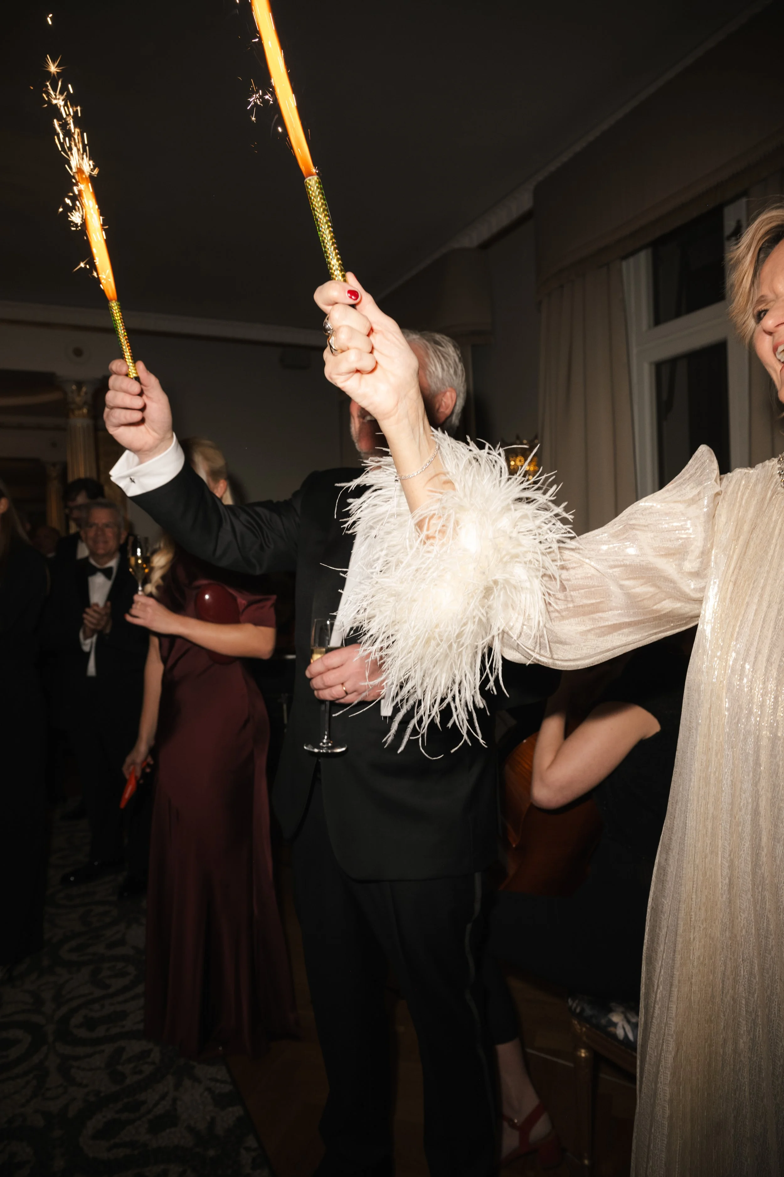 People celebrating at a party, holding sparklers and champagne glasses, dressed in formal attire, with some smiling and enjoying the event.