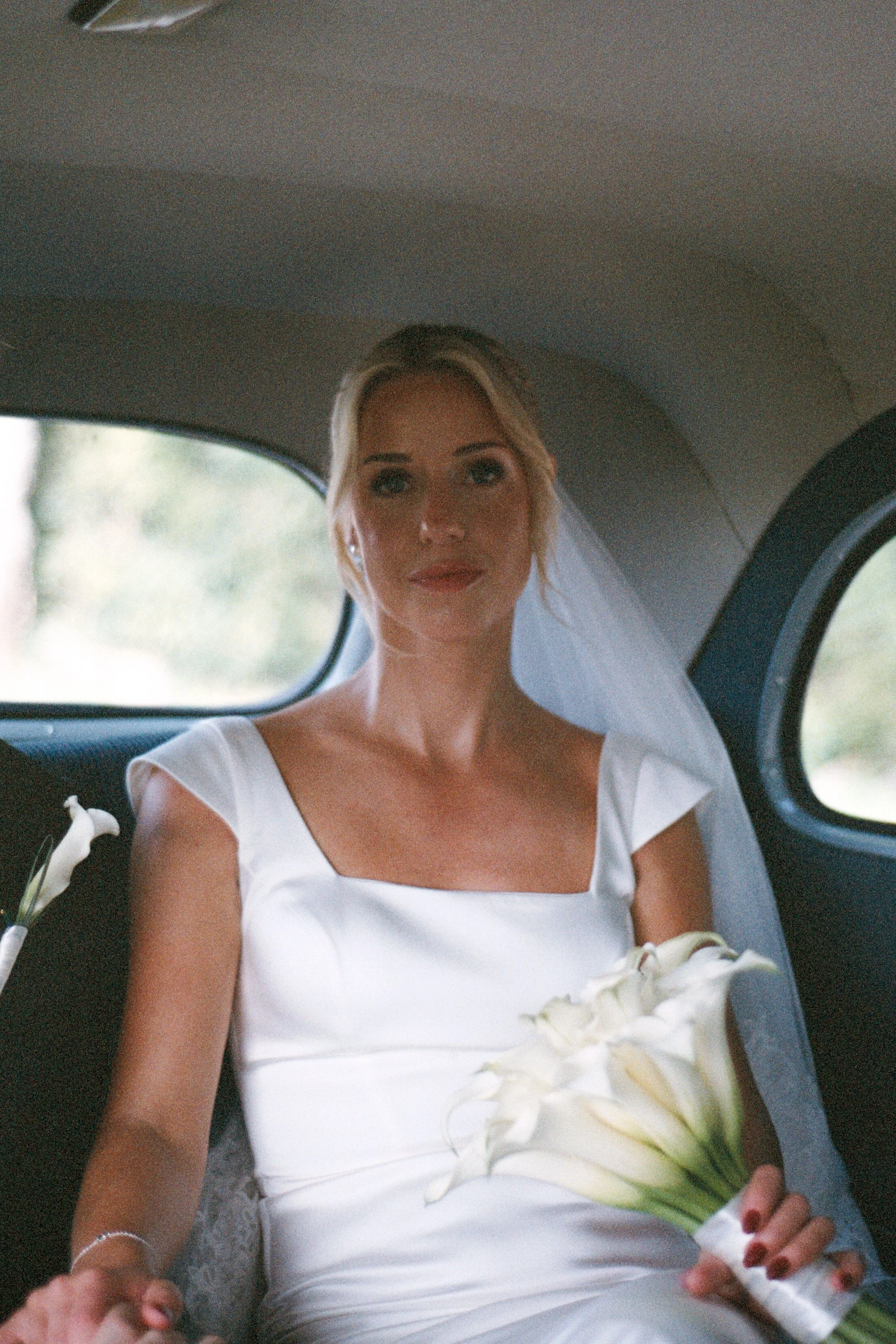 A bride with blonde hair dressed in a white wedding gown sitting inside a vehicle holding a bouquet of white calla lilies.