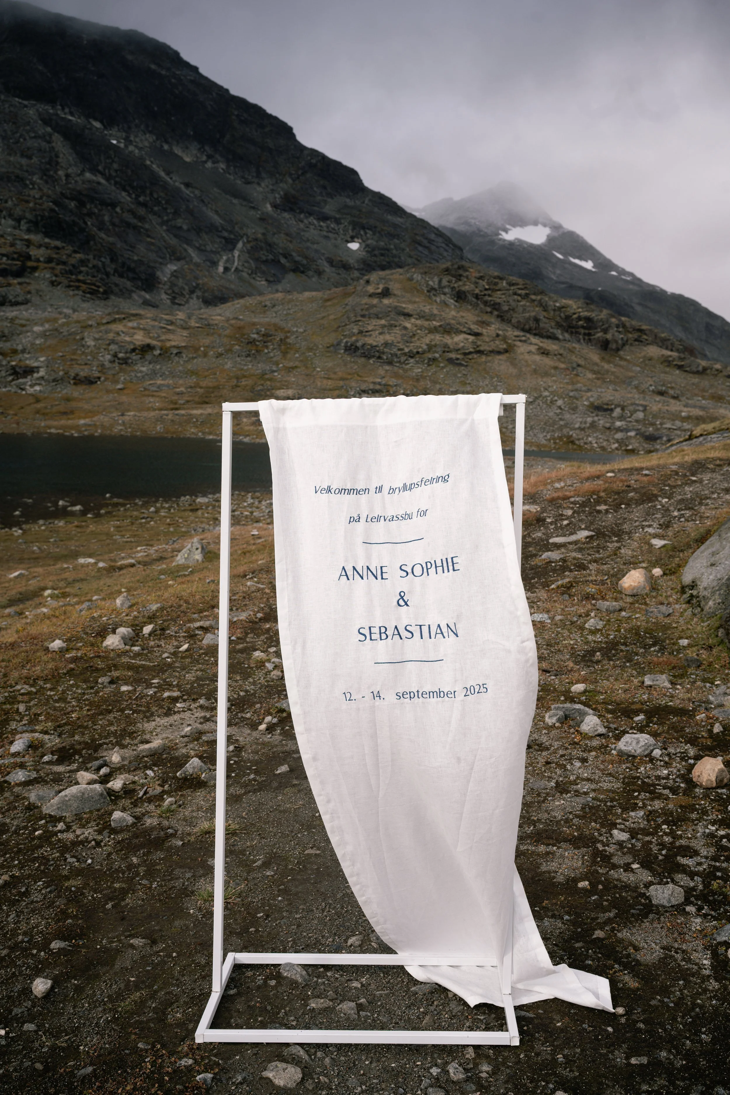 White fabric banner with wedding details on a metal stand outdoors in a mountainous landscape with rocky terrain, hills, and cloudy sky.